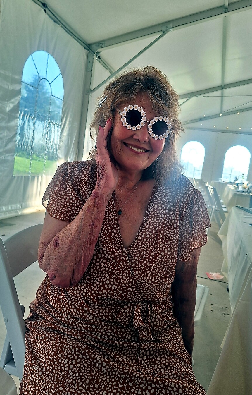 Smiling elderly woman in a brown patterned dress wearing oversized sunglasses with daisy frames inside a white event tent.
