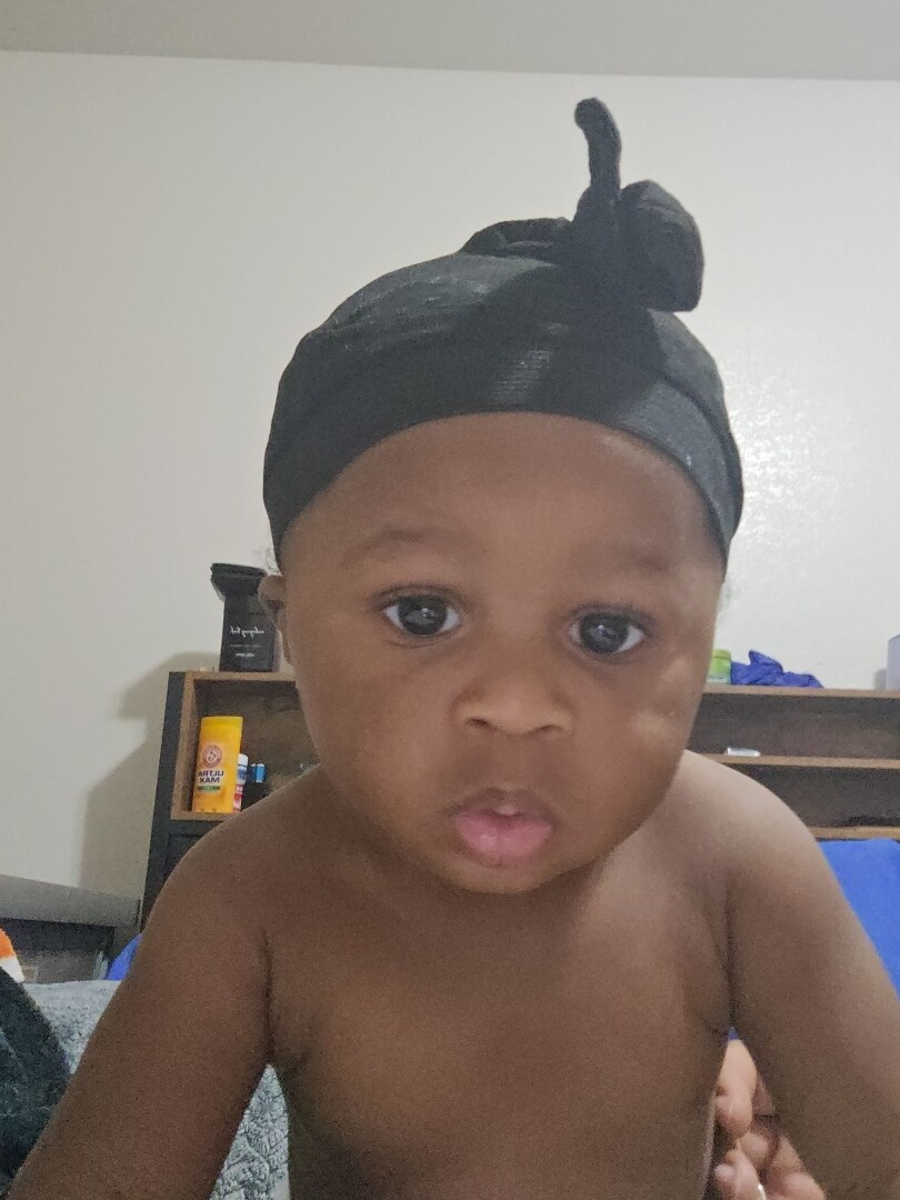 Infant with a black head wrap, shirtless, looking at the camera with a curious expression in a room with shelves behind him.