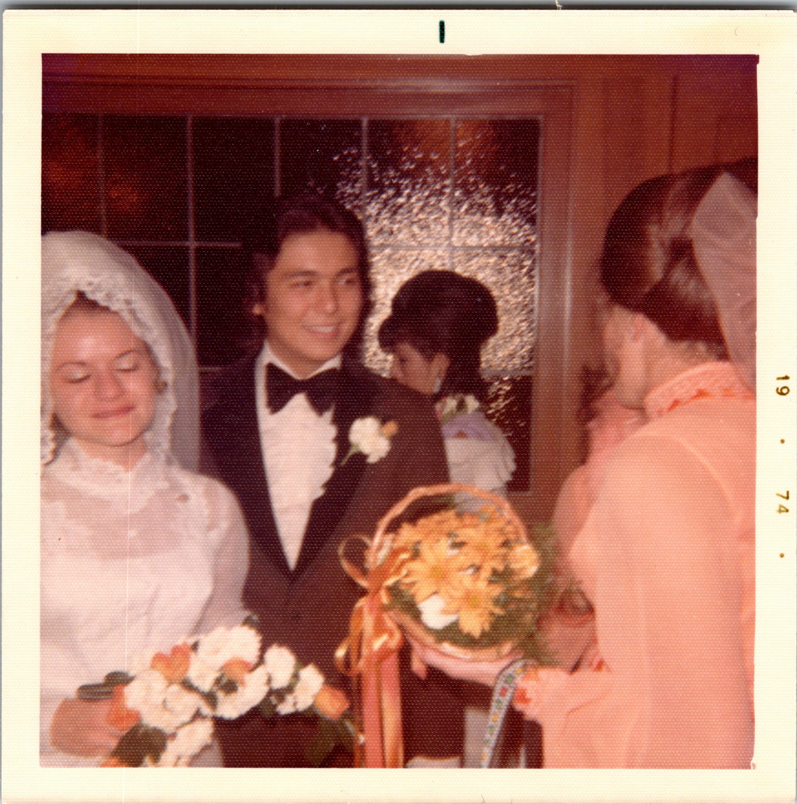 Group photo at a wedding: guests in formal attire, a man in a tuxedo with boutonniere, women in dresses including a hijab, and a floral bouquet visible.