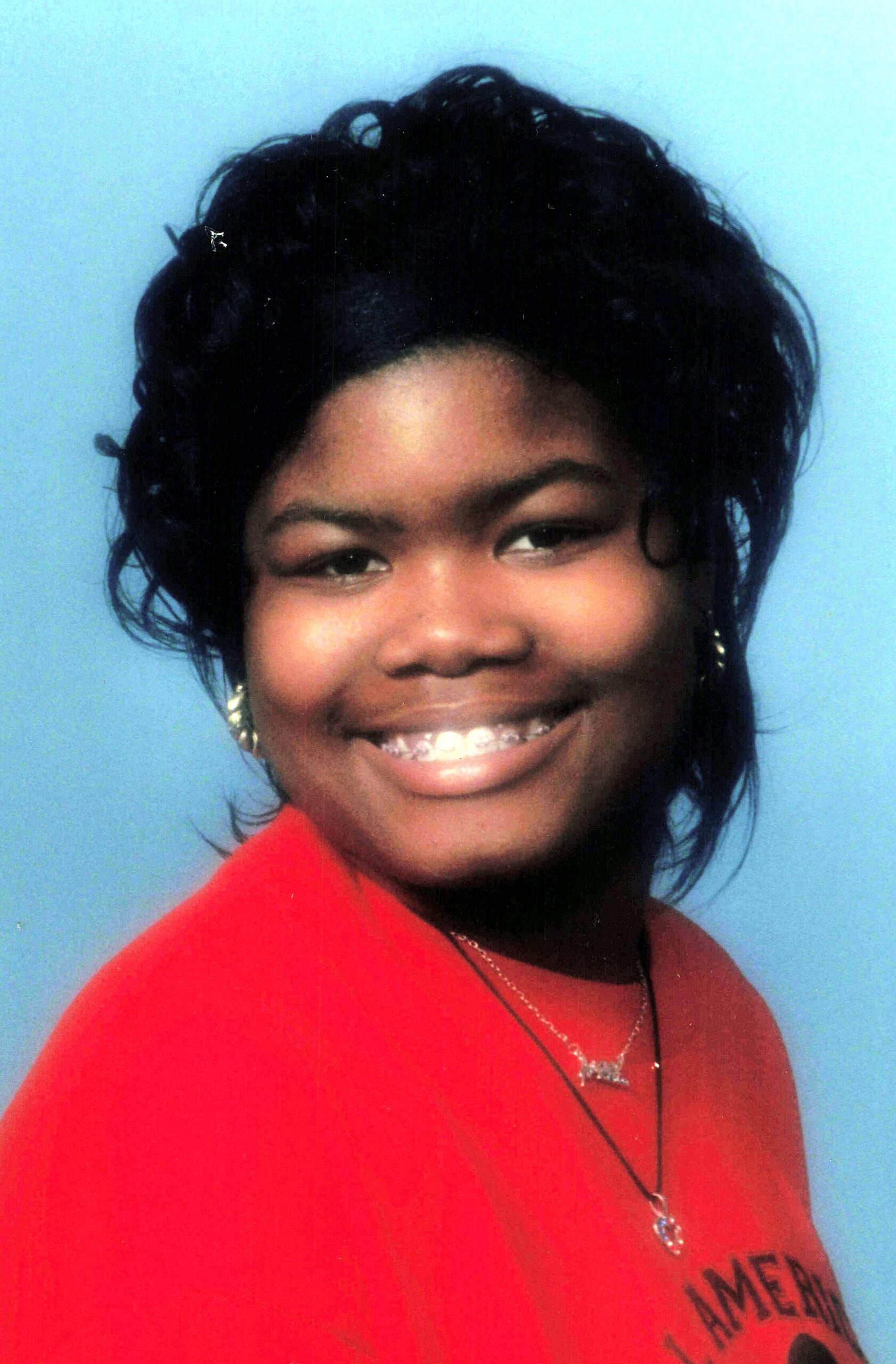 Smiling Black woman with short black hair wearing a red top against a light blue background, showing braces on her teeth.