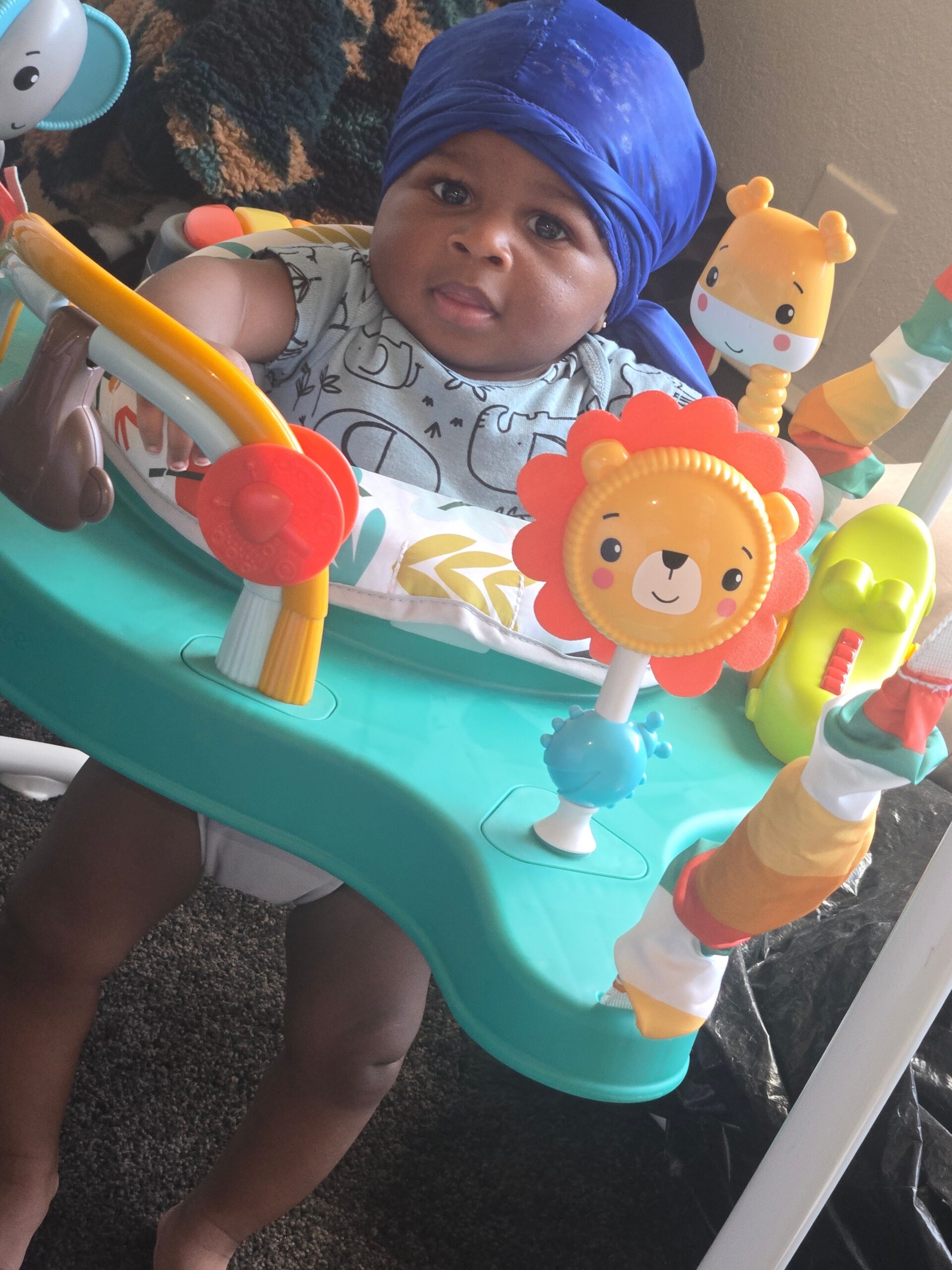 Infant in a turquoise activity jumper wearing a blue headwrap, surrounded by colorful toys including a smiling flower toy.