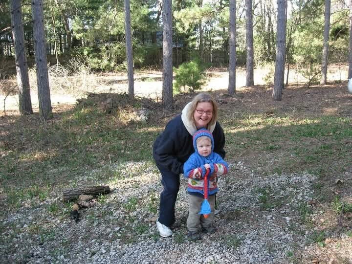 Woman bending beside a toddler outdoors in a wooded area, smiling at the camera; autumn light filtering through trees behind them.