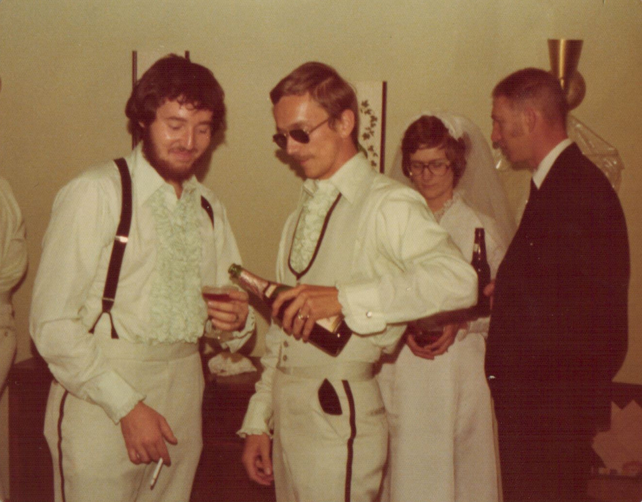 Two men in white 1970s-style wedding outfits pour champagne at a reception, with a bride and groom in the background.