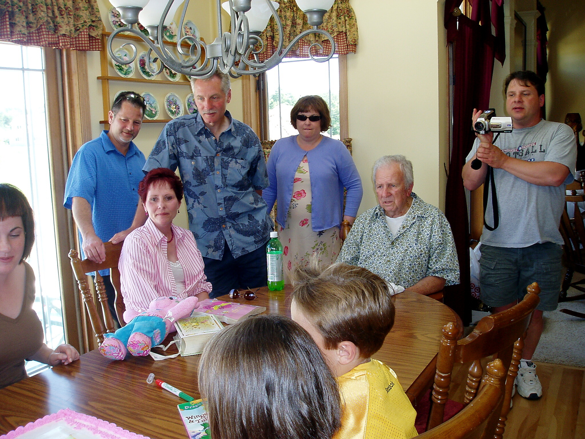 Family gathering around a wooden dining table in a sunlit room; a man on the right records with a video camera while others sit or stand nearby.
