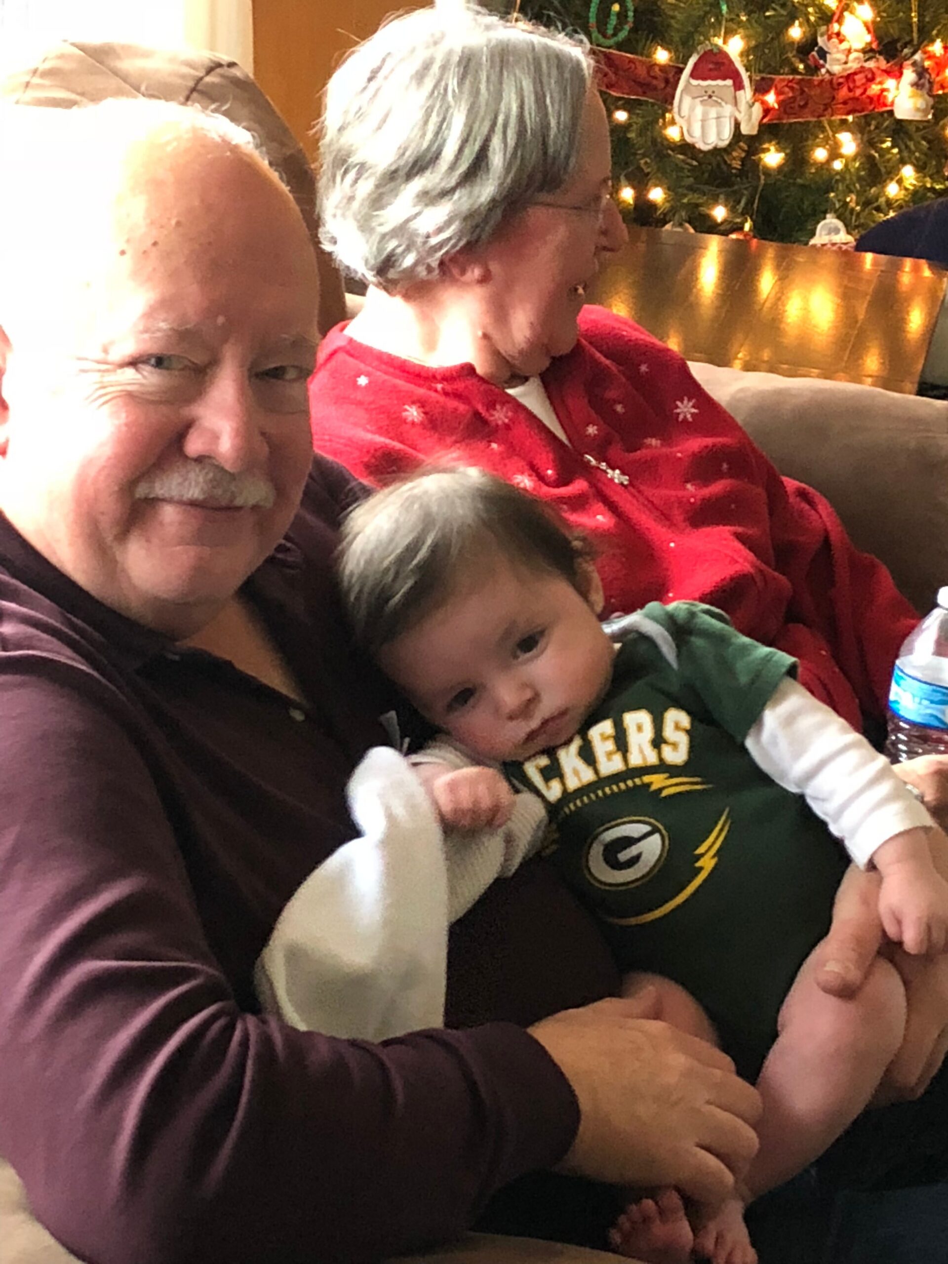 Family portrait on a couch during Christmas: an older man with a mustache holds a baby in a Packers onesie while a woman in a red sweater sits beside them, a Christmas tree lit with lights in the background.