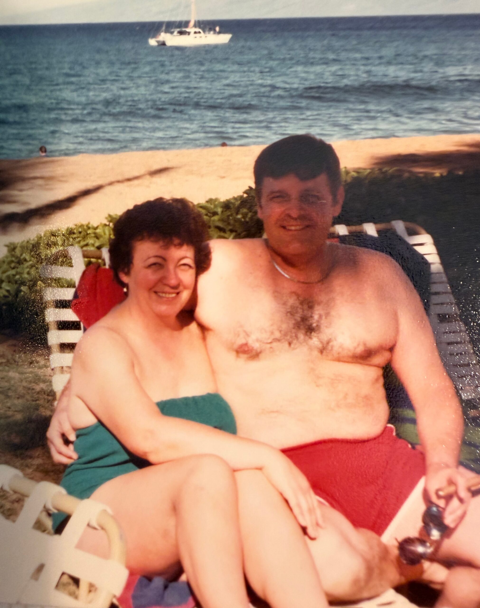 Smiling couple lounging on a beach chair by the sea, with a sailboat in the distance.