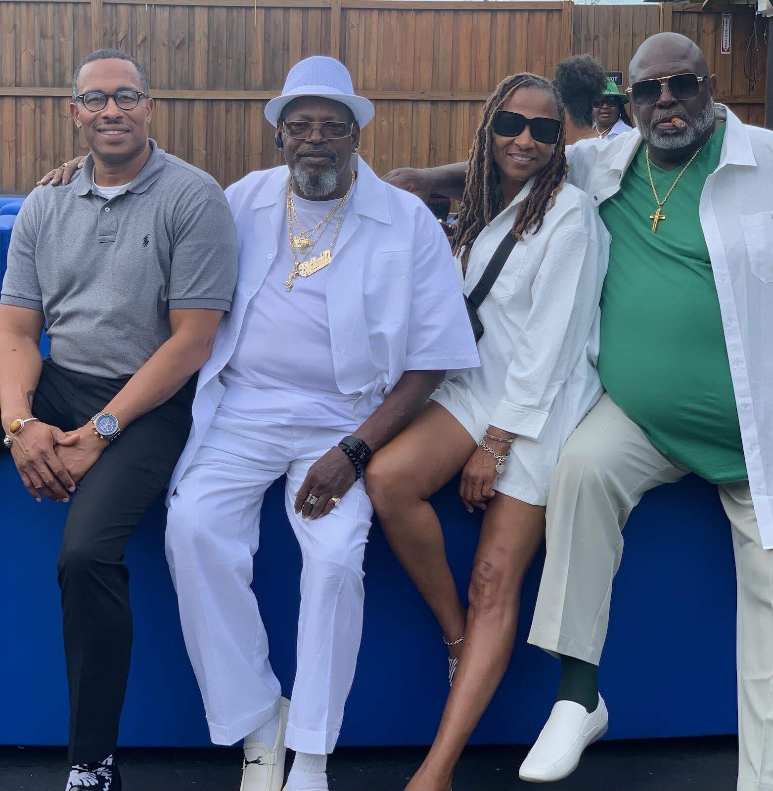 Four adults posing together outdoors, seated along a blue barrier with a wooden fence behind them. From left to right: a man in a gray polo, a man in white with hat and gold jewelry, a woman with dreadlocks and sunglasses, and a man in green shirt and white jacket wearing a gold necklace and cross.