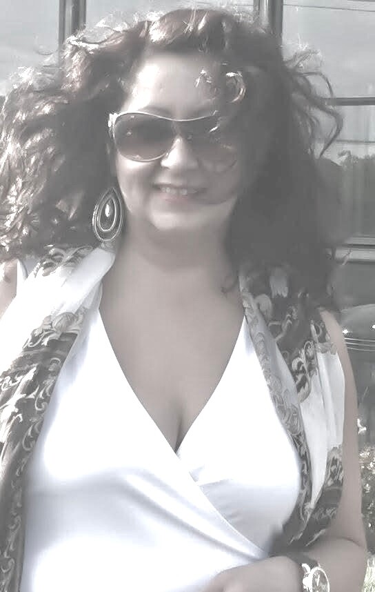 Smiling woman outdoors in daylight wearing a white top, patterned scarf, large sunglasses, and hoop earrings.