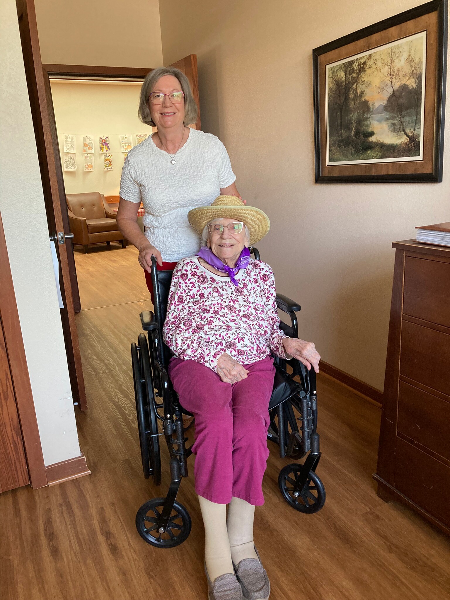 Two women in a home hallway: an elderly woman in a wheelchair wearing a hat, floral top, purple pants, and beige tights, with a smiling caregiver standing beside her in a white textured top.