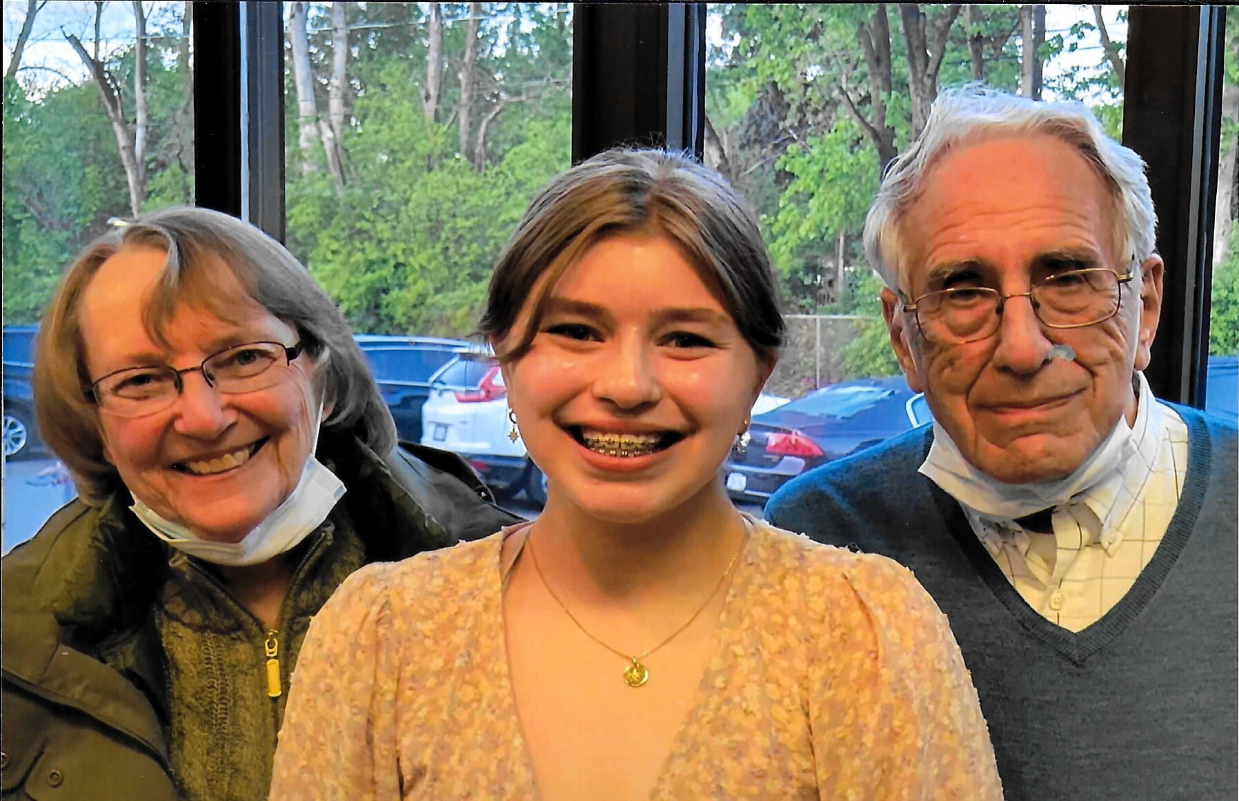 Three people posing for a photo: an smiling older woman on the left, a braces-wearing girl in the center, and an older man on the right, with masks pulled under their chins.