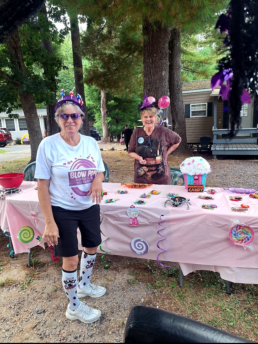 Two women at a candy-themed outdoor party, one in a white T-shirt and knee-high socks standing by a pink decorated table.