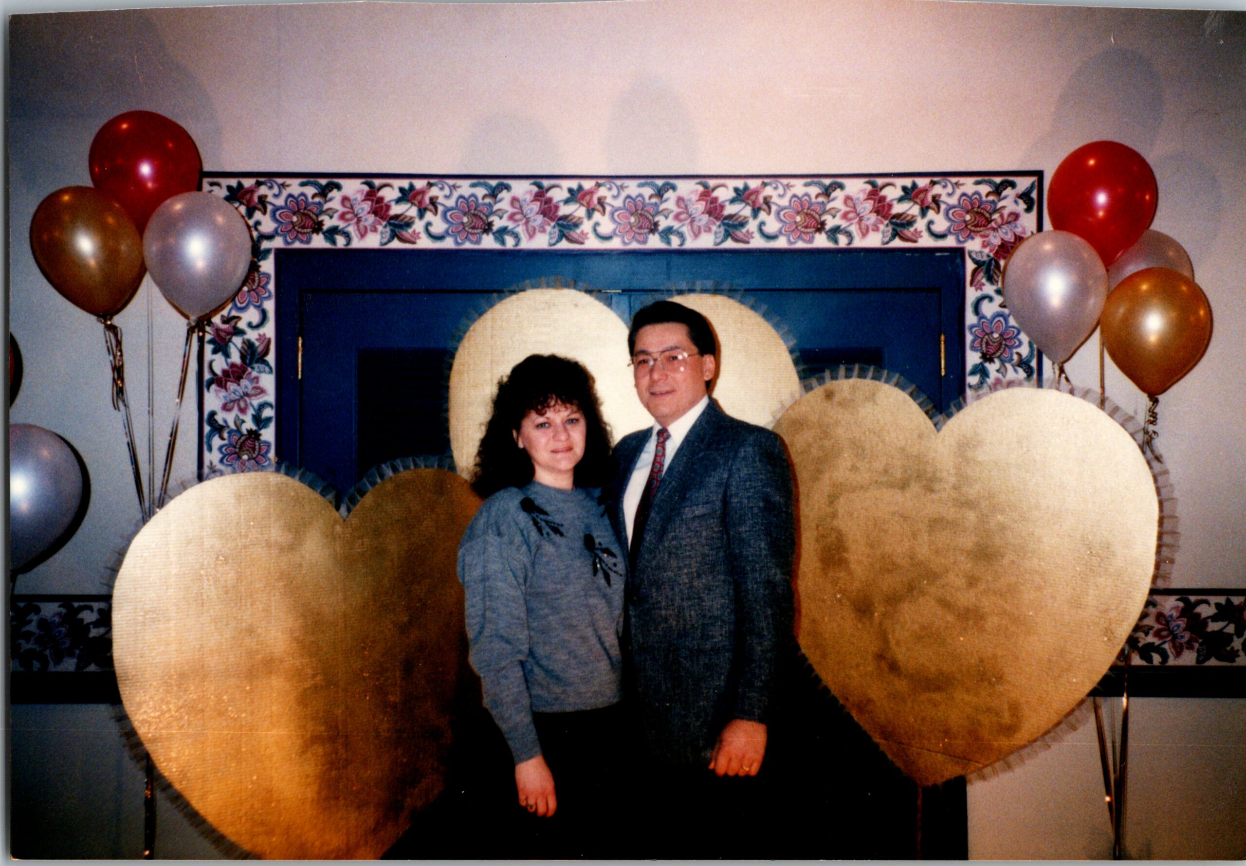 Couple posing at a celebration, framed by oversized gold paper hearts and balloons on both sides.
