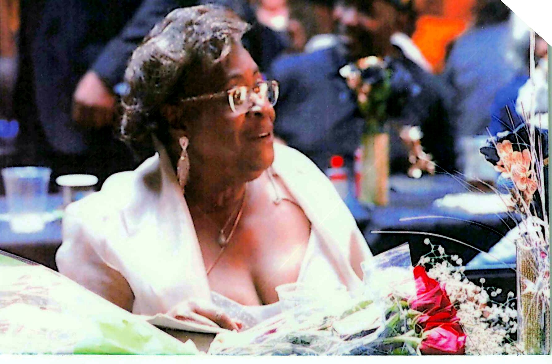 Elderly woman in a light outfit and glasses gazes toward a bouquet of flowers at a memorial.