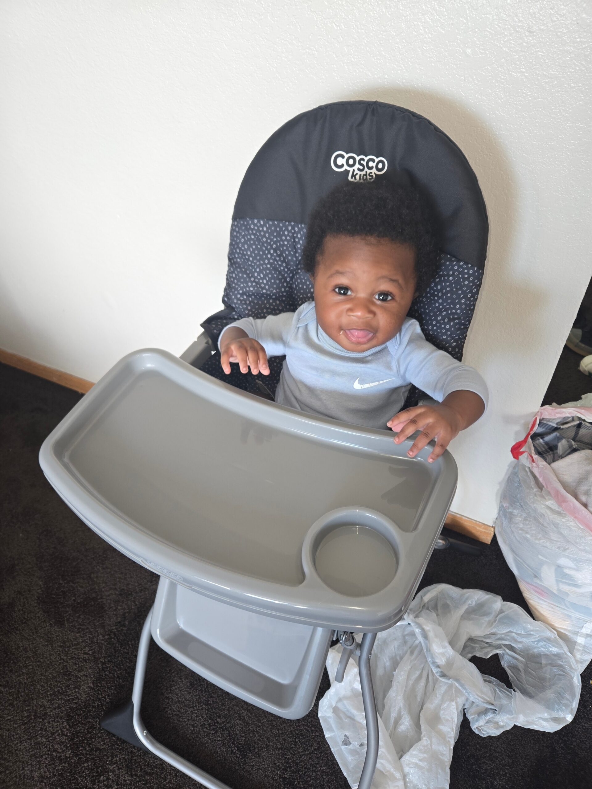 Baby sitting in a gray Cosco Kids high chair with a detachable tray, smiling at the camera.