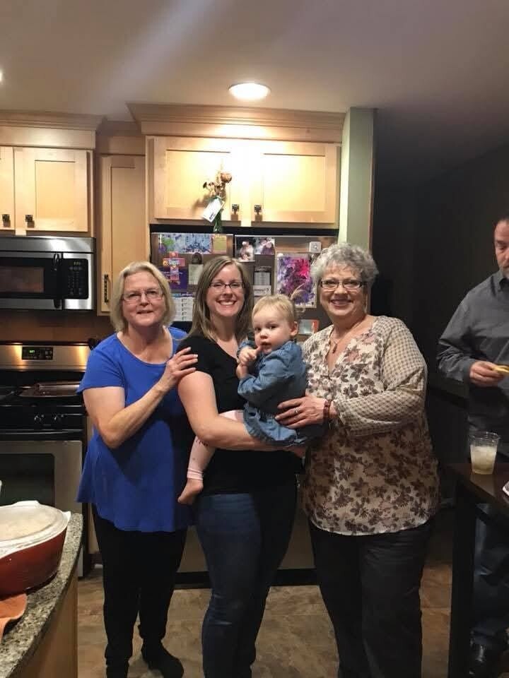 Group of four adults in a kitchen posing for a photo, with a baby held by one woman, all smiling at the camera.
