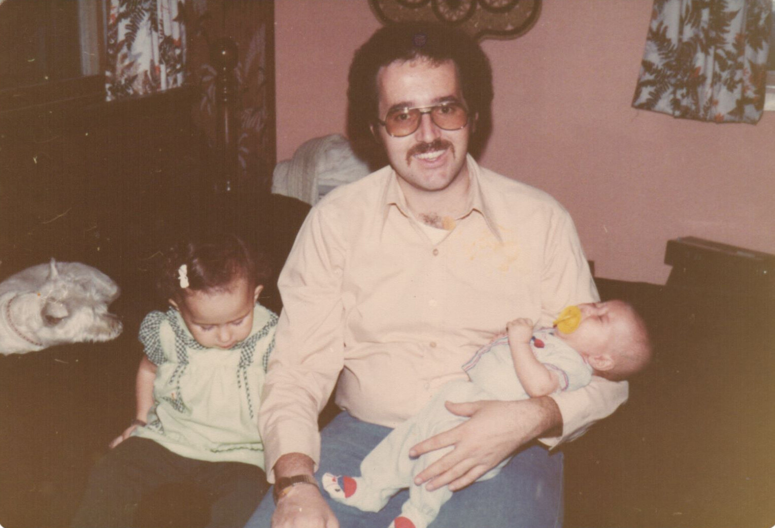 Adult man with glasses and a mustache sits on a couch, holding a baby who is sucking a pacifier while a young girl sits beside him and a small dog rests nearby in a pink-walled living room.