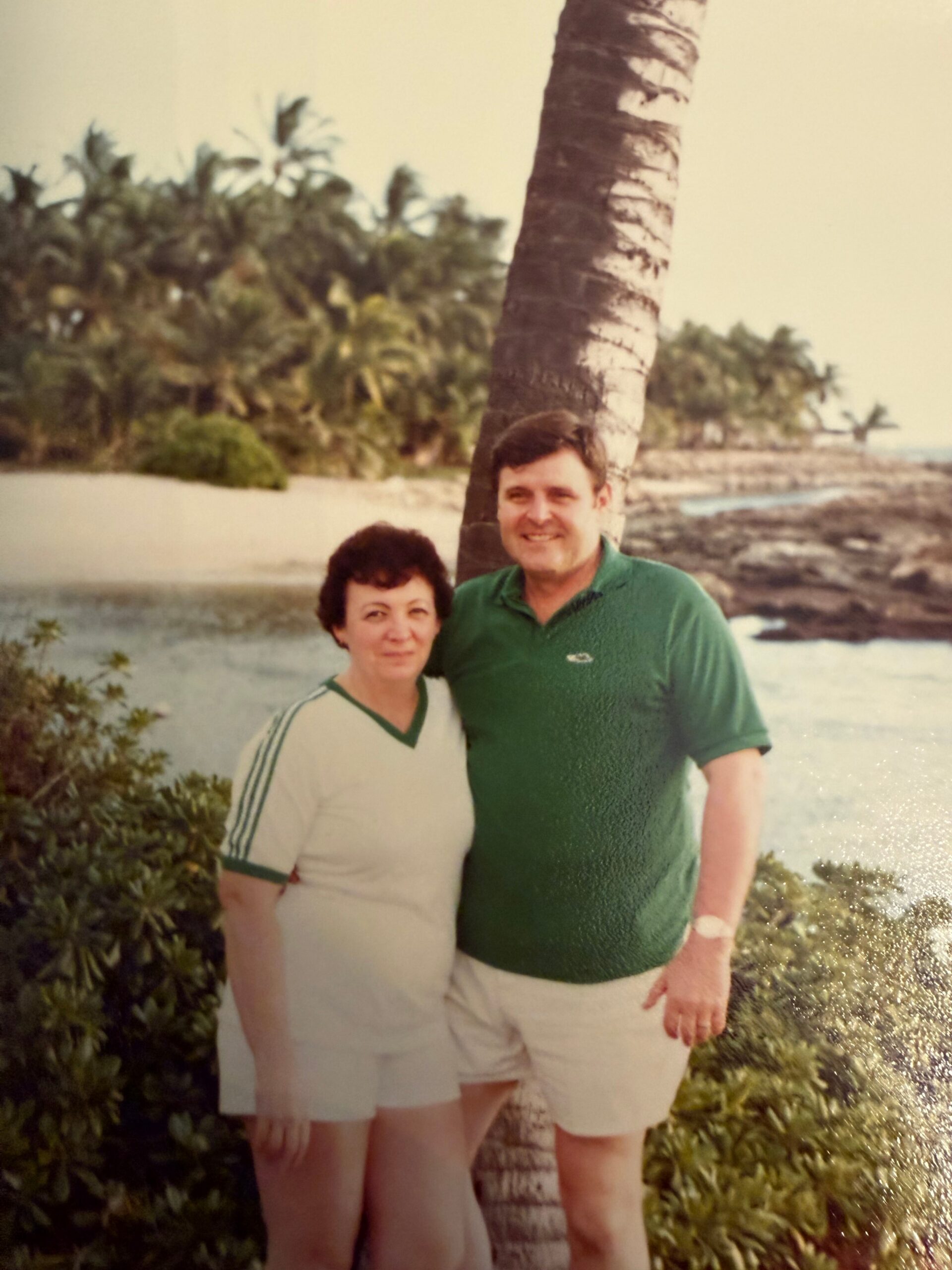 A couple posing together by a palm tree on a sunny beach with greenery and water in the background, smiling at the camera.
