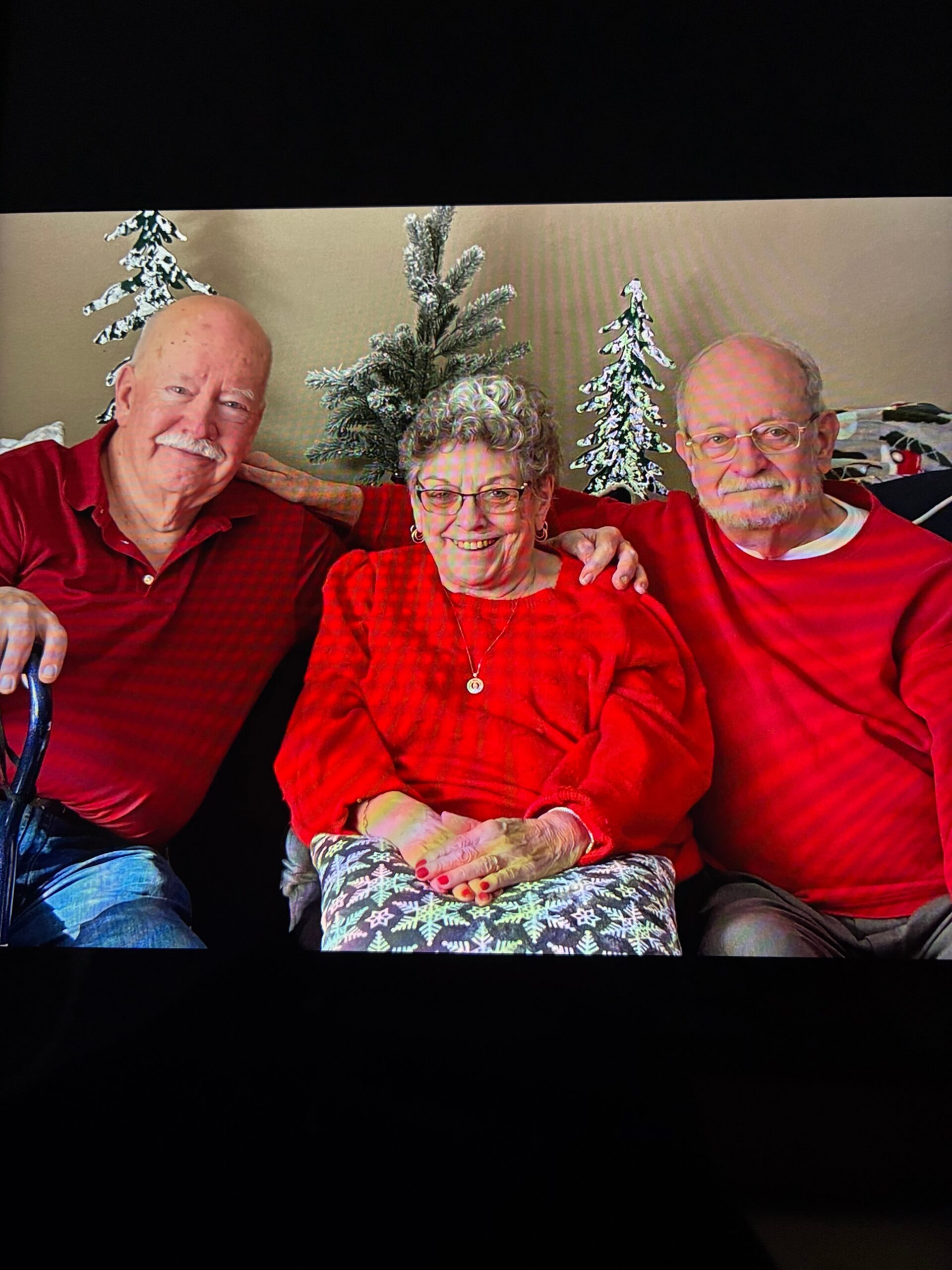Three smiling seniors in red outfits sit close together on a sofa, with snowy miniature Christmas trees in the background behind them.