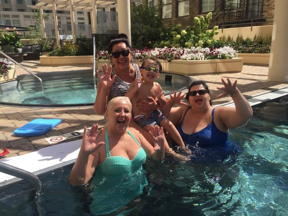 Four adults and a child in a rooftop pool, smiling and waving at the camera on a sunny day.