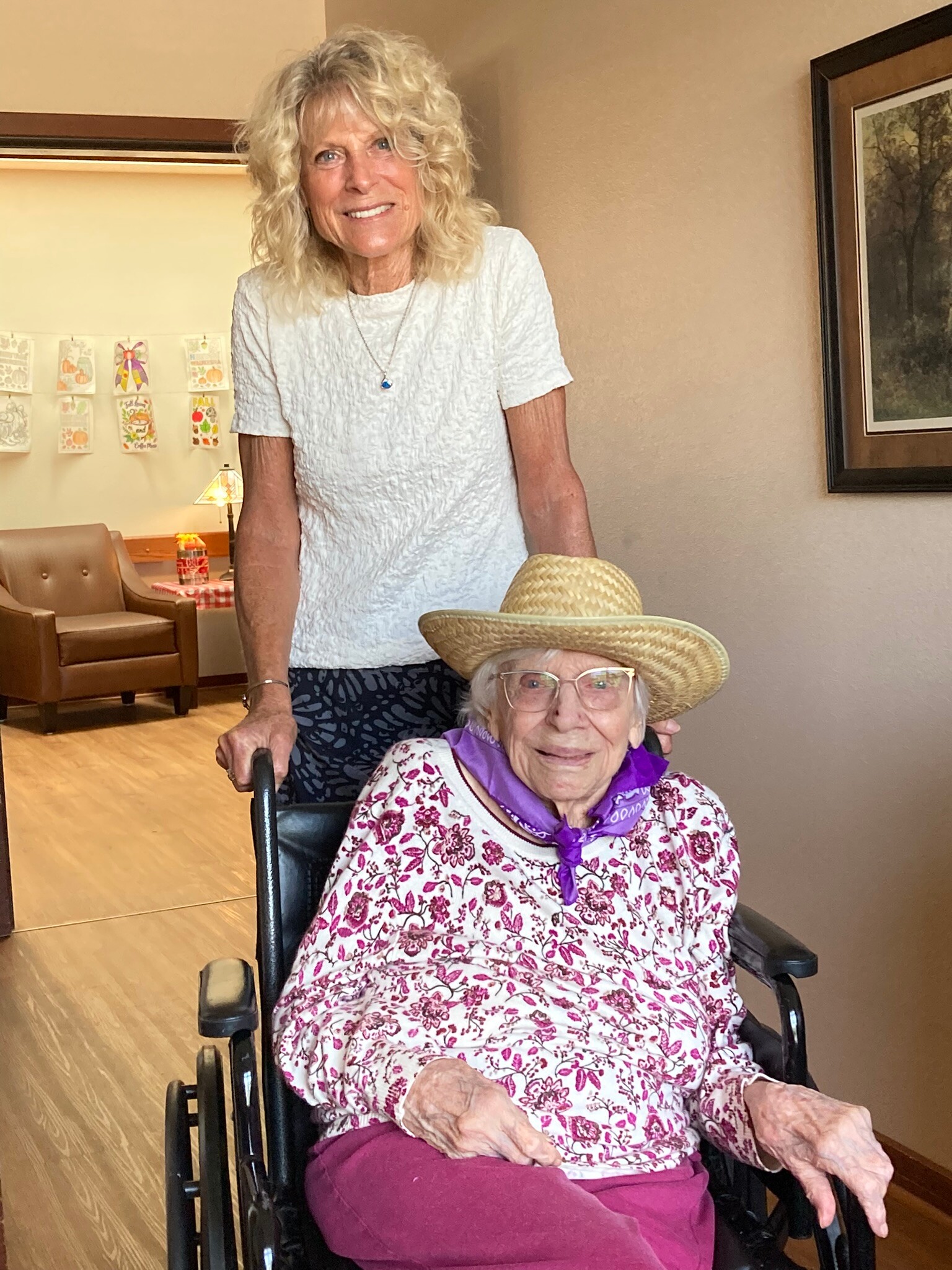Smiling elderly woman in a floral blouse sits in a wheelchair while another woman stands behind her in a white top in a cozy room.