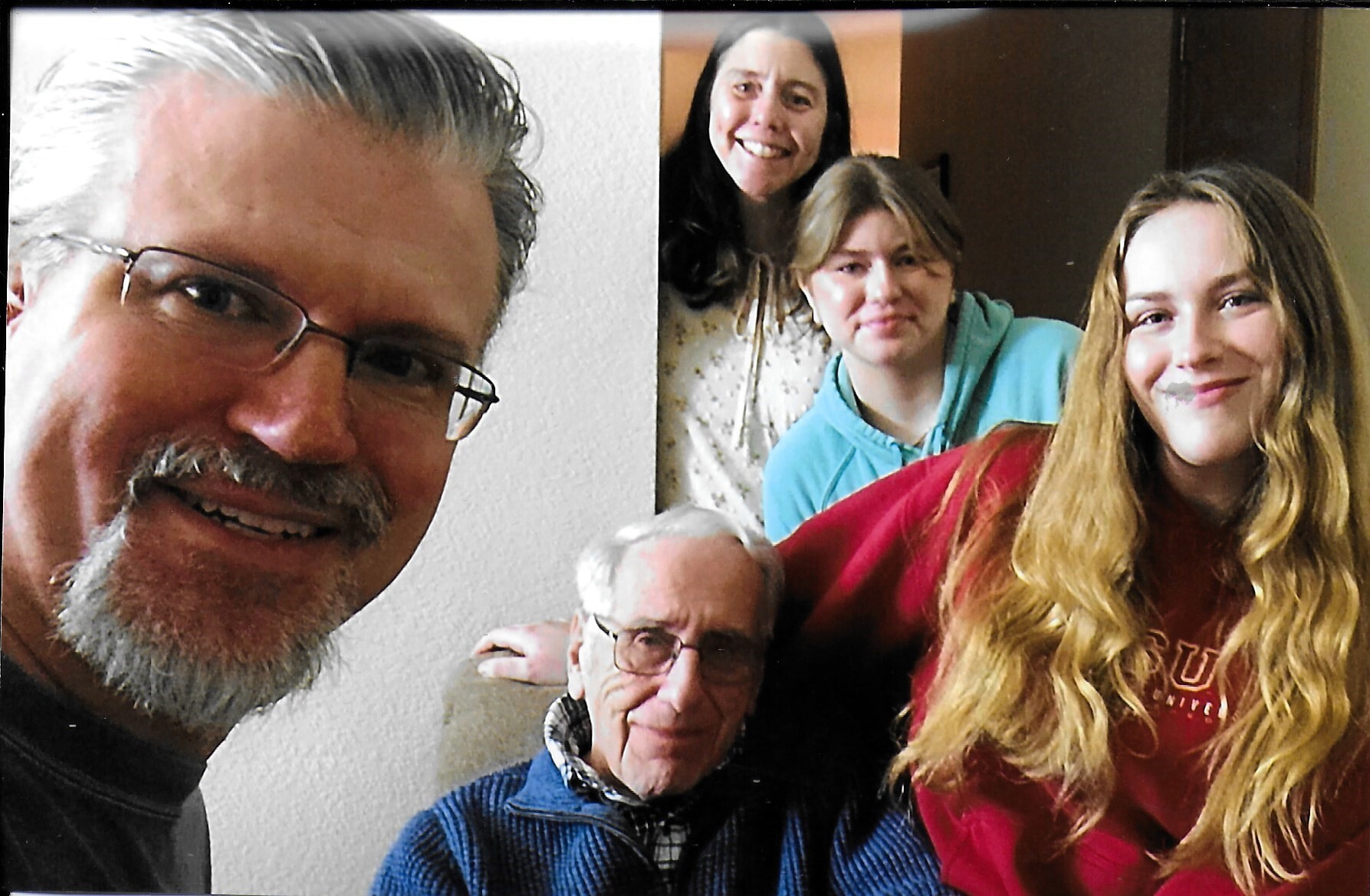 Close-up group selfie of five smiling people: man with glasses on the left and four others in a living room background.