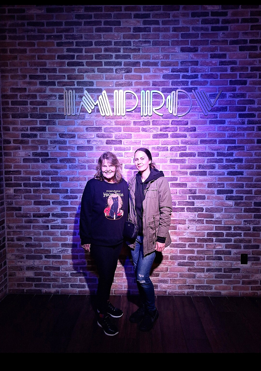 Two women posing and smiling in front of a brick wall with a purple neon sign overhead.