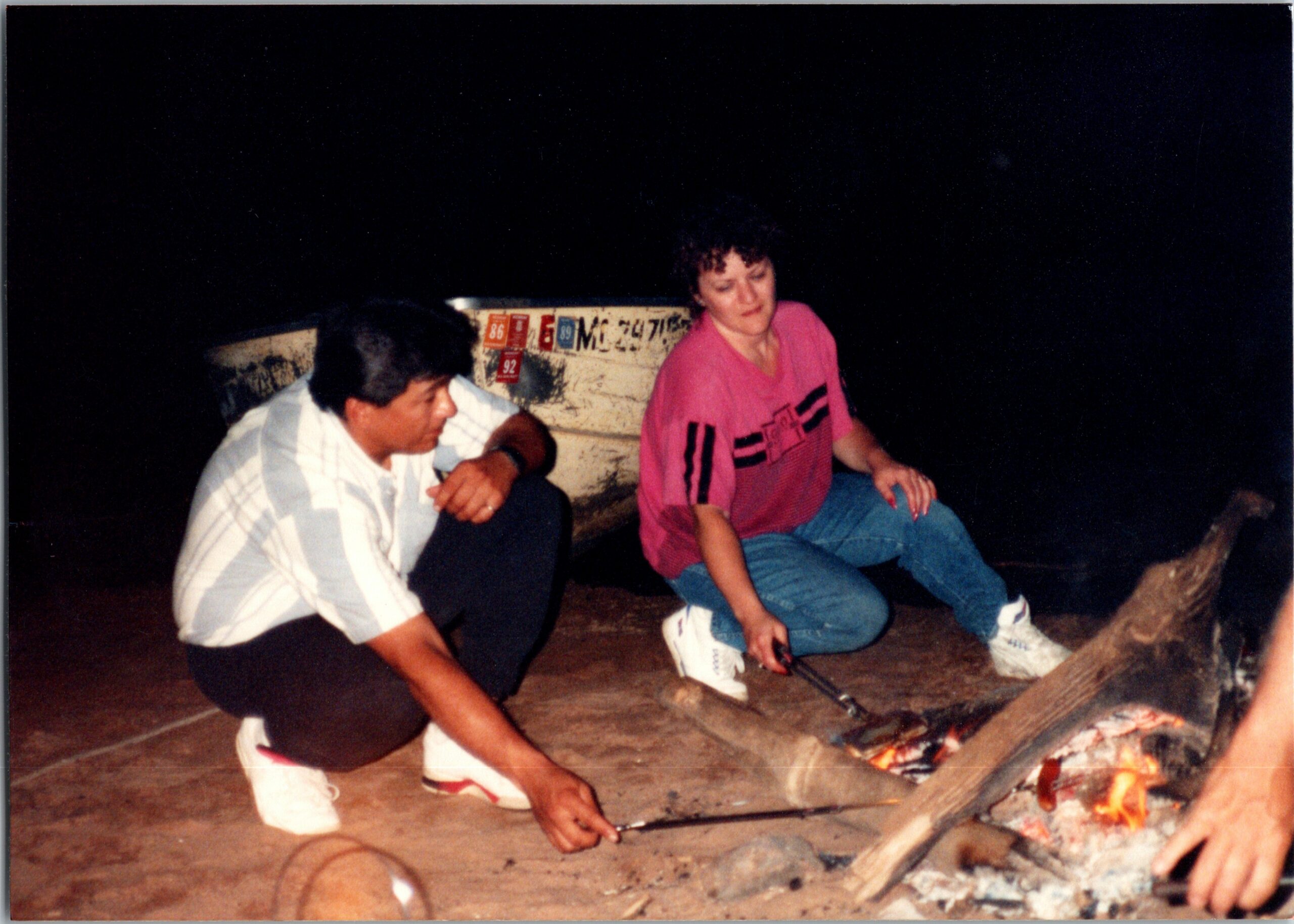 Two people crouched near a glowing campfire or grill outdoors at night, tending meat on skewers or a spit.