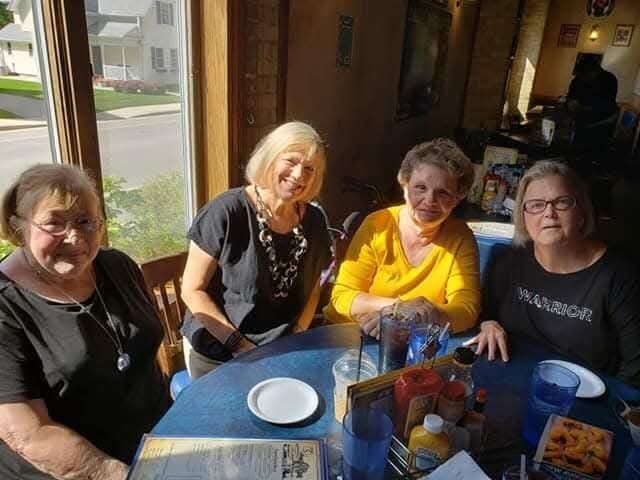 Four women sit around a blue table in a sunlit restaurant, smiling and chatting together at a window table.