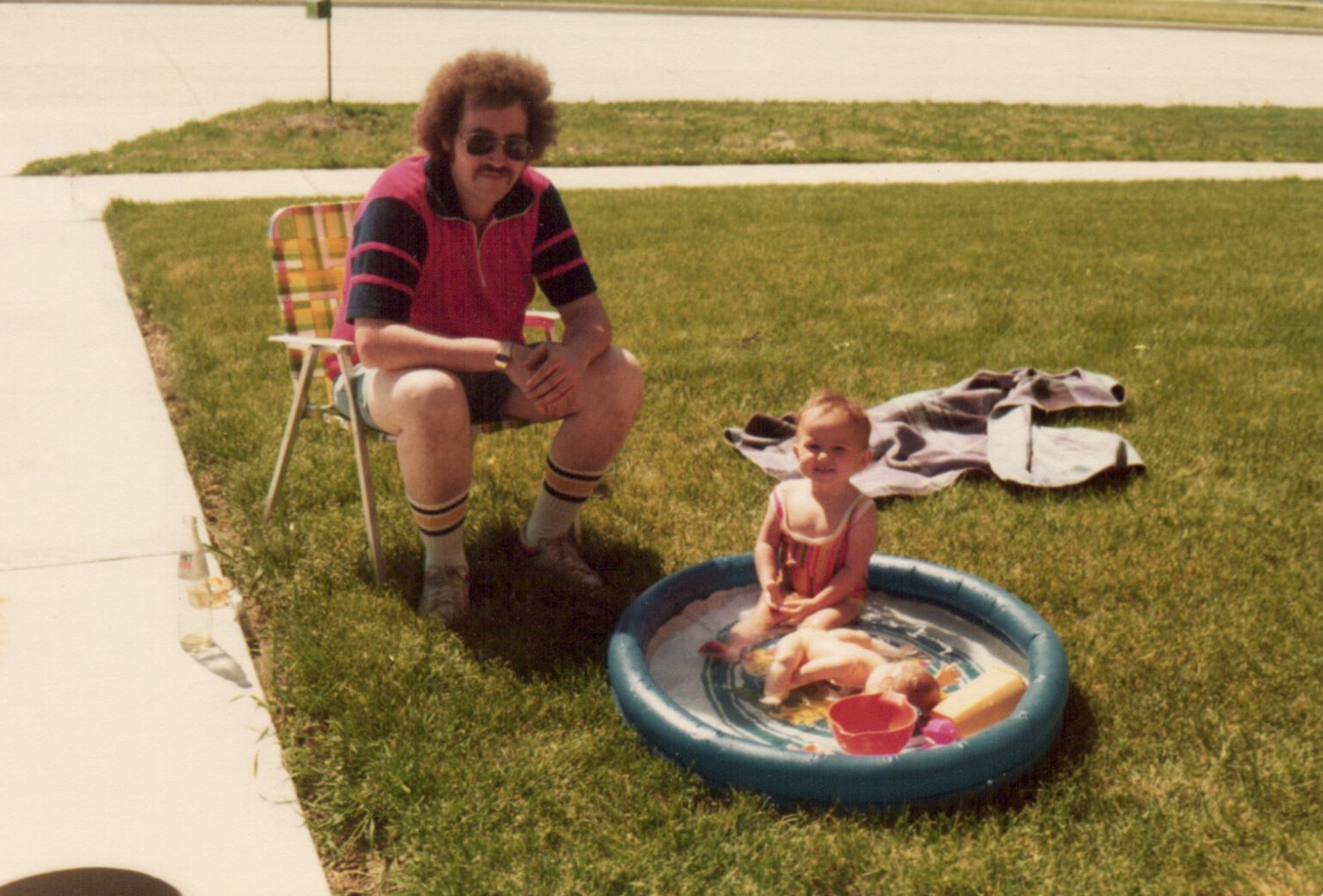 Man with curly hair and sunglasses sits in a plaid lawn chair watching a toddler in a small blue kiddie pool on a sunny suburban lawn.