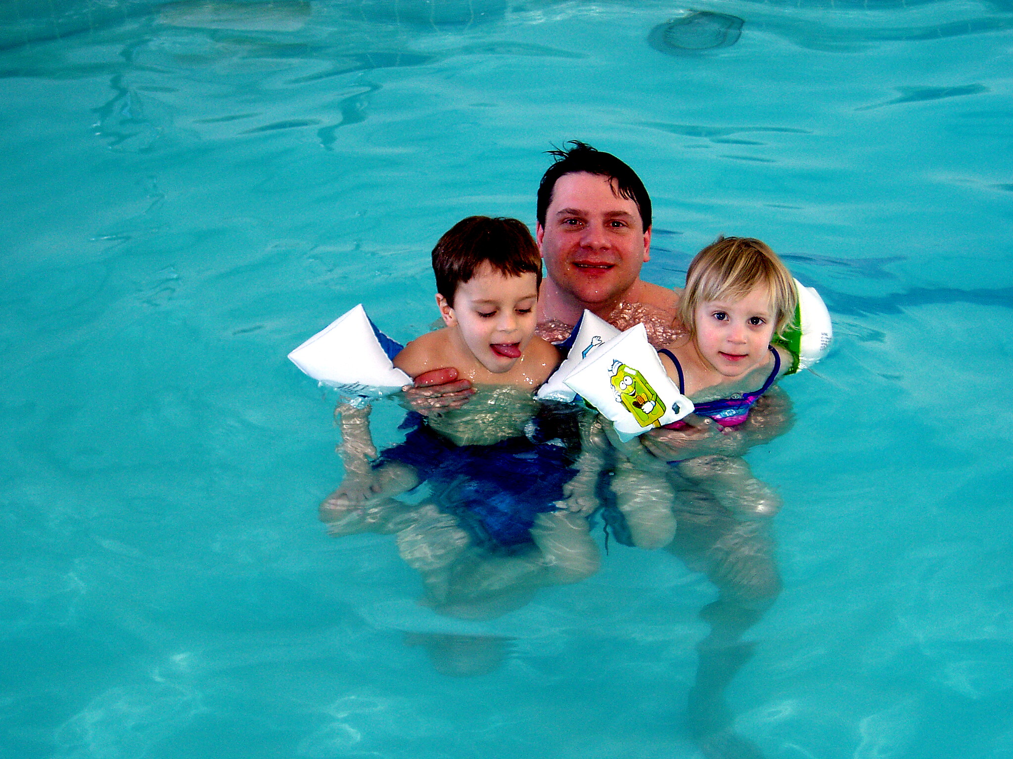 Adult with two children swimming in a bright blue pool, wearing inflatable arm floats with cartoon designs.