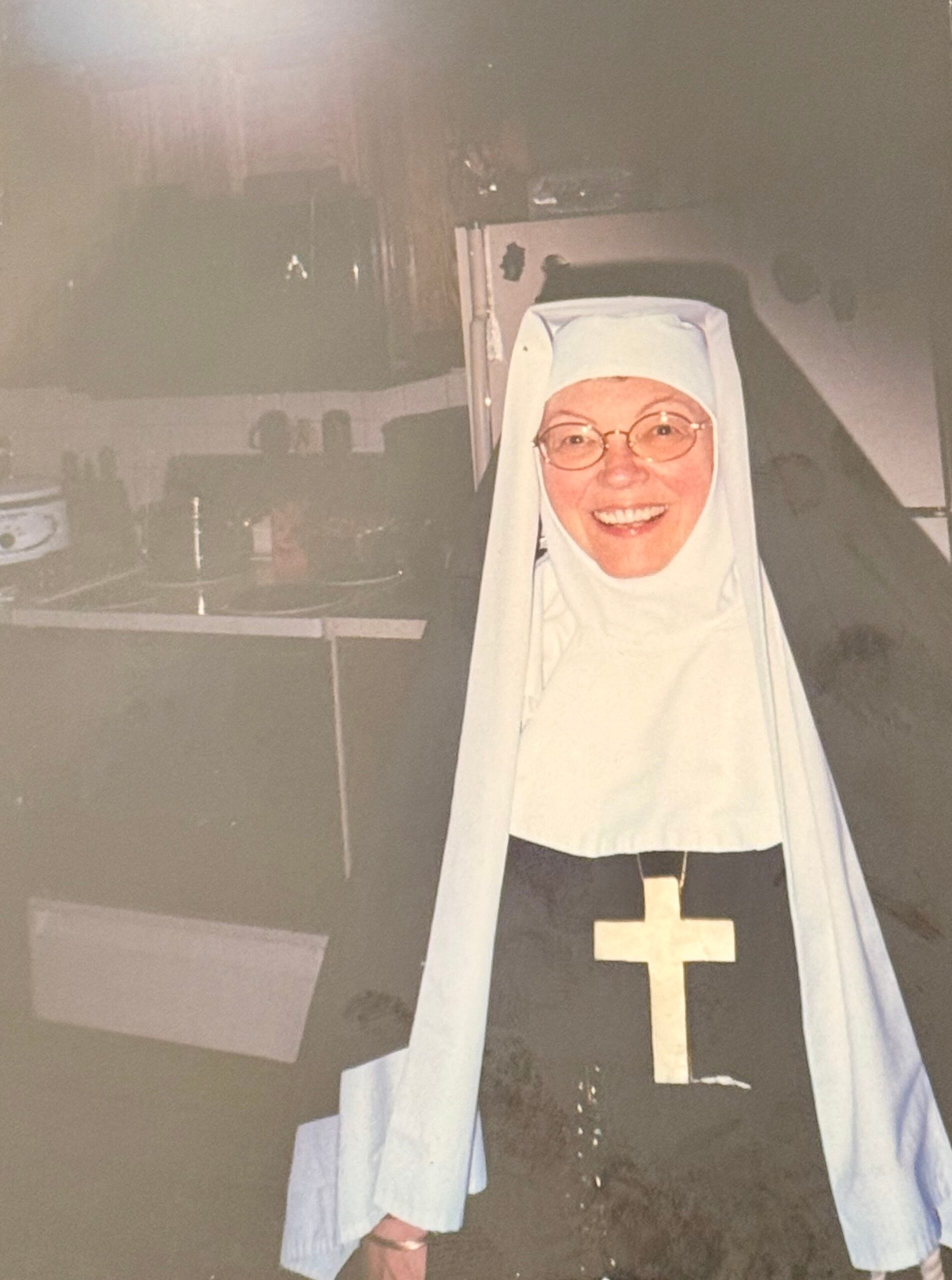 Smiling nun wearing a light blue veil and white habit with a gold cross on the front, indoors in a church or chapel setting.