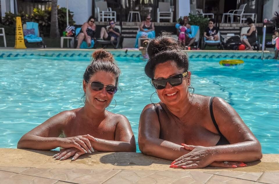 Two women in sunglasses lean on the pool edge, smiling at the camera as a sunny resort pool and loungers shimmer in the background.