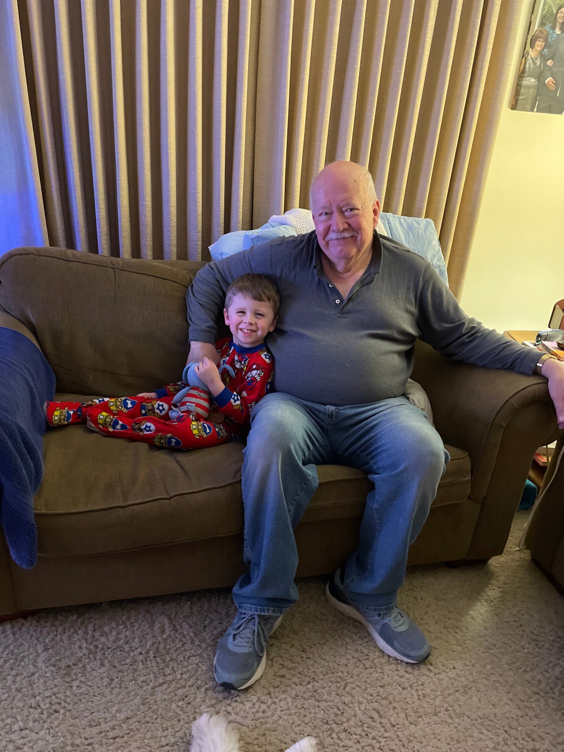 Grandfather and young boy sit together on a brown sofa, smiling at the camera, with beige curtains behind them. The child wears red superhero pajamas.