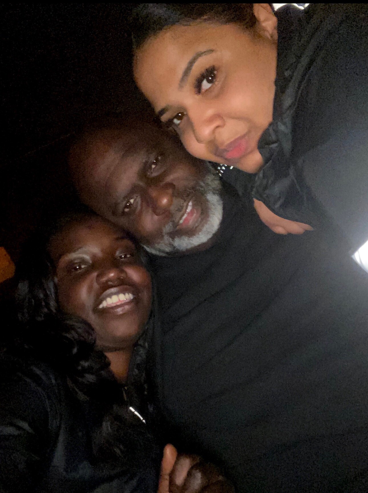 Three friends taking a close-up selfie at night, smiling for the camera on a dark background.