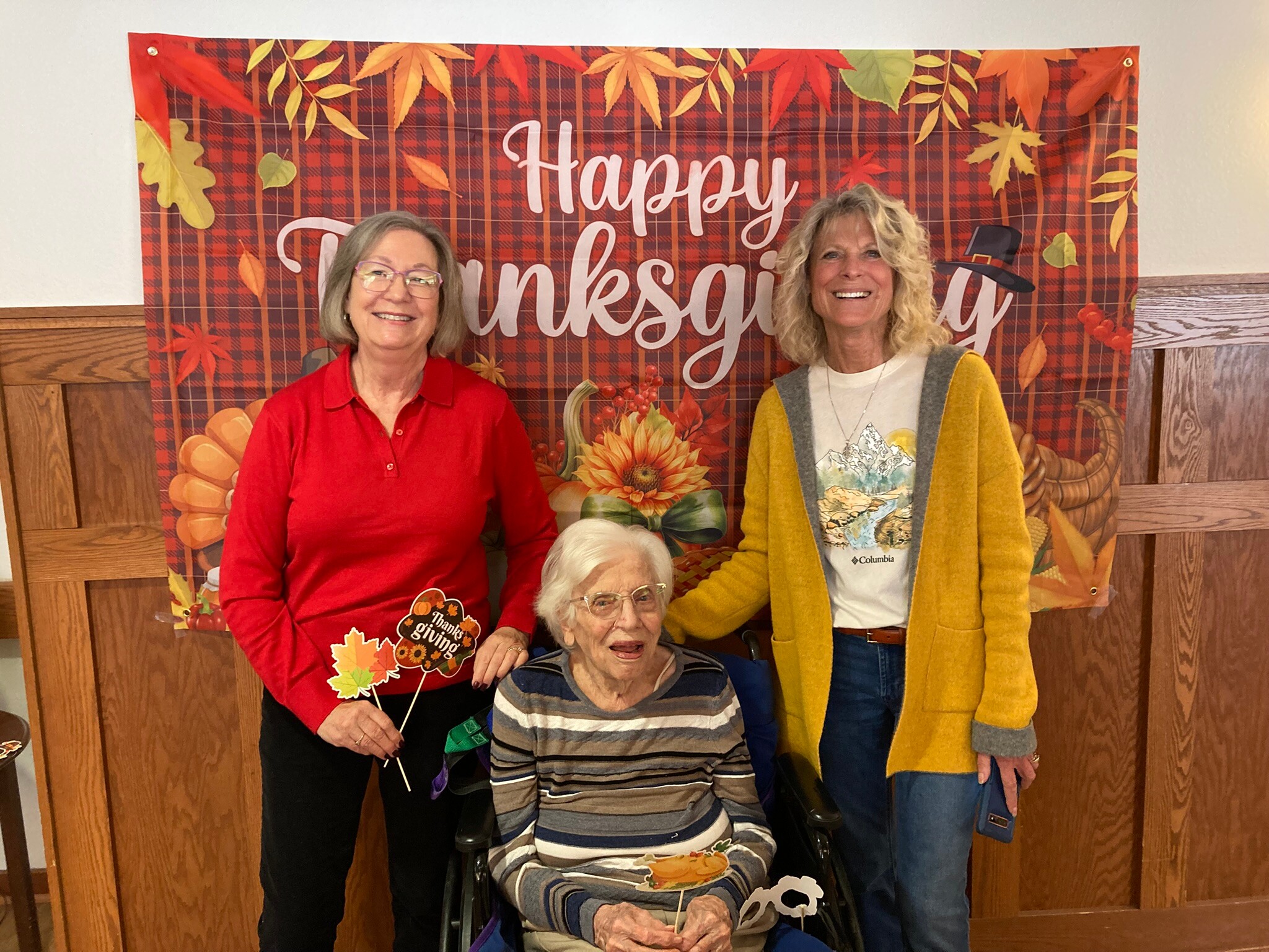 Group of four people posing for a Thanksgiving photo in front of a autumn-themed banner with leaves and pumpkins. A woman in red stands left, a woman in a yellow cardigan stands right, and a seated elderly woman holds fall props.