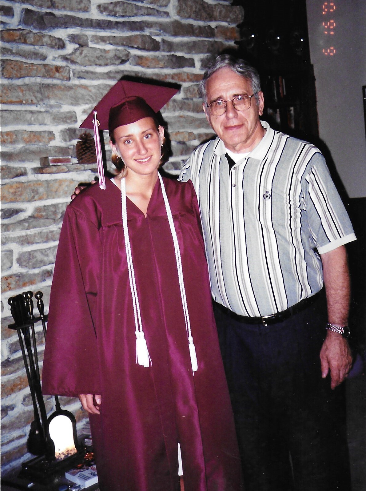 Young woman in a maroon graduation gown and cap with white cords beside an older man in a striped shirt, standing by a stone fireplace indoors.