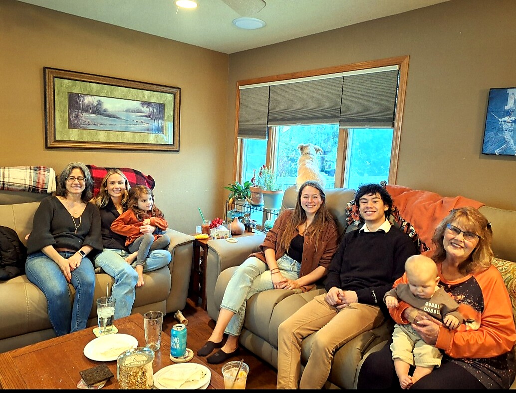 Family portrait in a cozy living room: six adults and two young children sit on couches, a dog by the window behind them, snacks on the coffee table.