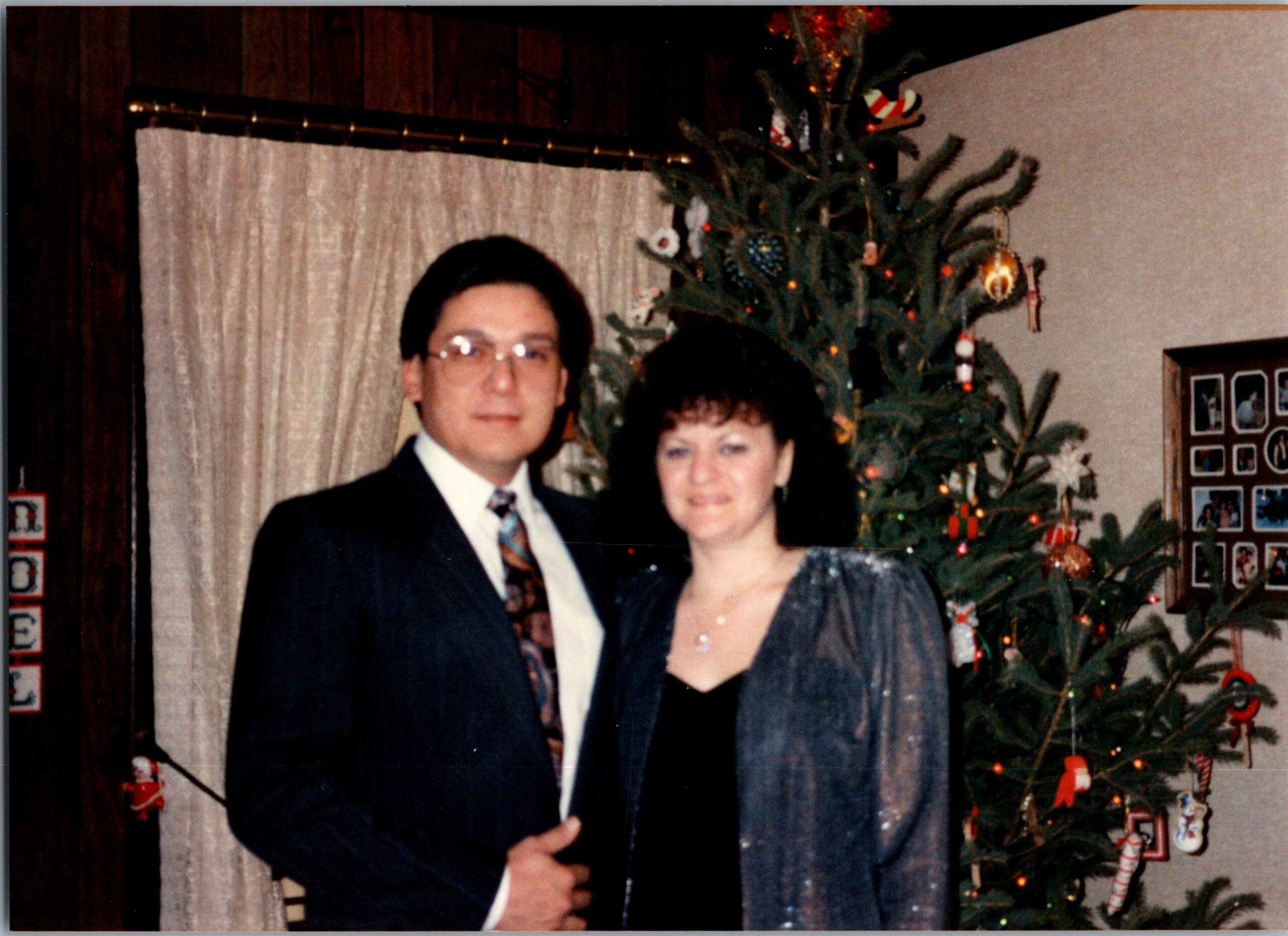 Man in a suit and woman in a dress standing with their arms around in front of a decorated Christmas tree indoors, smiling at the camera.
