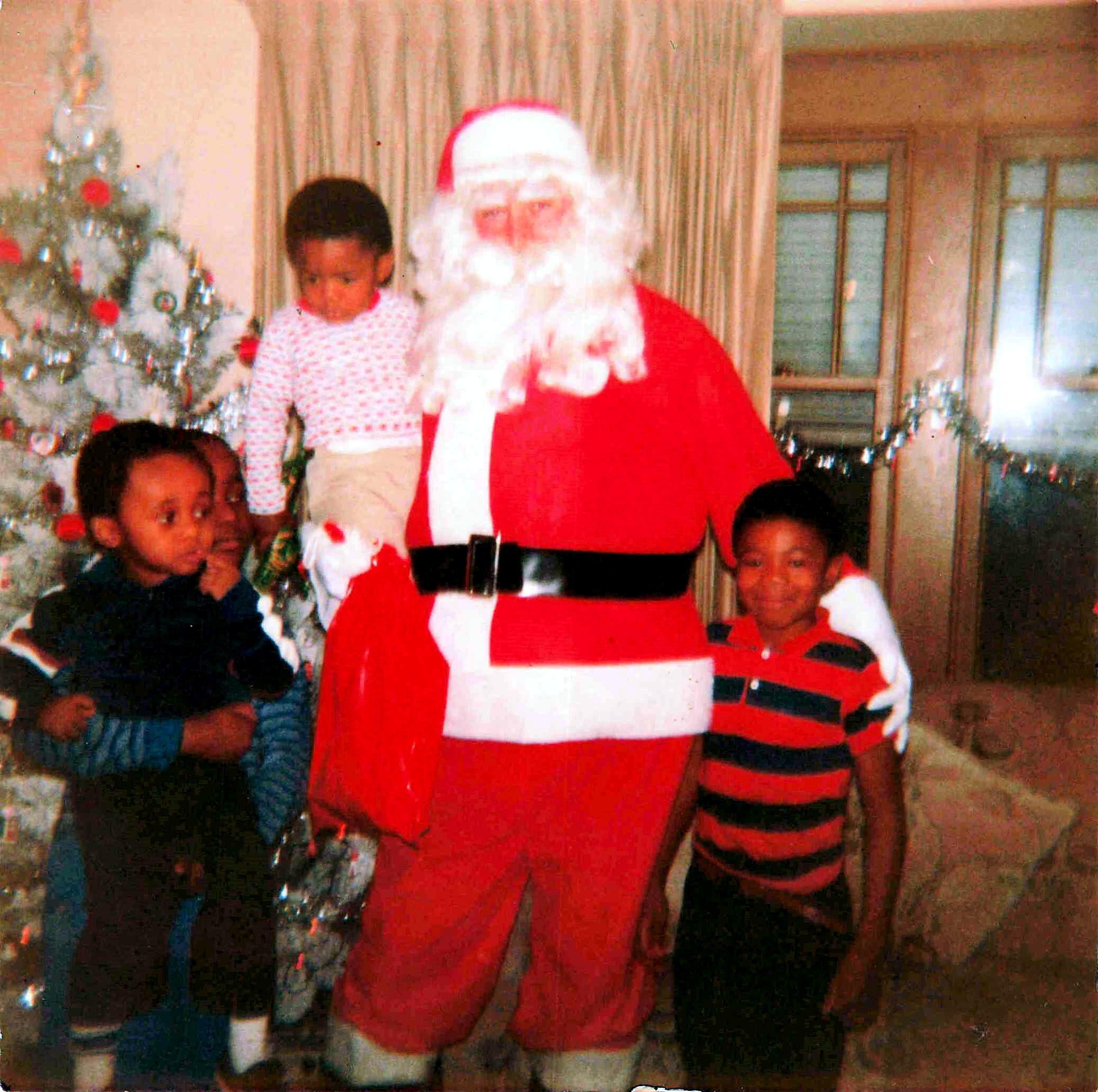 Santa Claus in a red suit standing with children indoors by a Christmas tree during the holidays.
