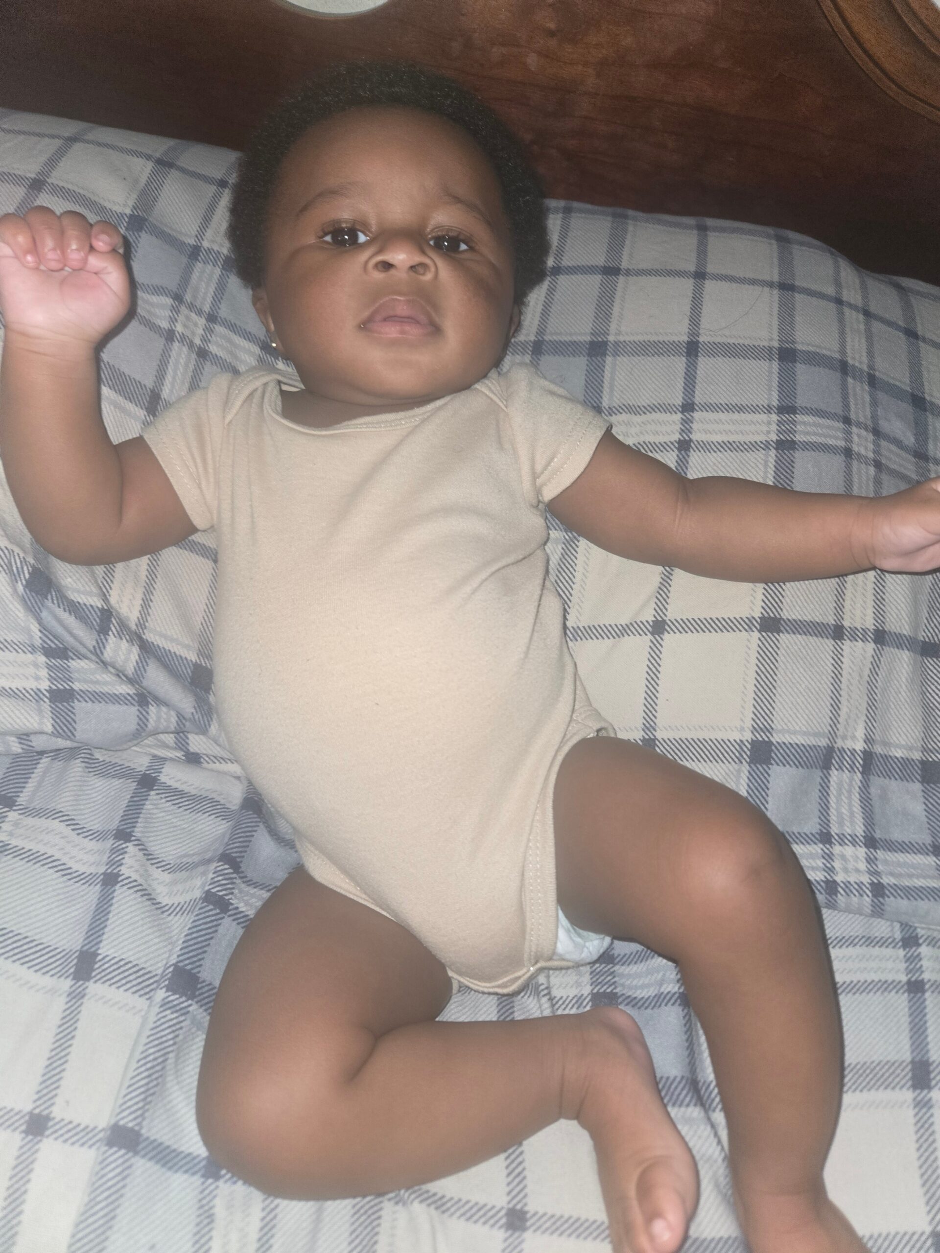 Infant lying on a plaid blanket with arms raised, wearing a beige onesie, looking at the camera.