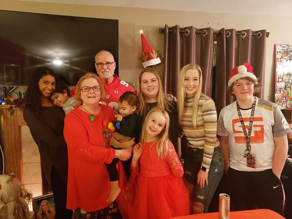 Group of family members posing indoors for a festive holiday photo, some wearing Santa hats and red outfits, smiling at the camera.