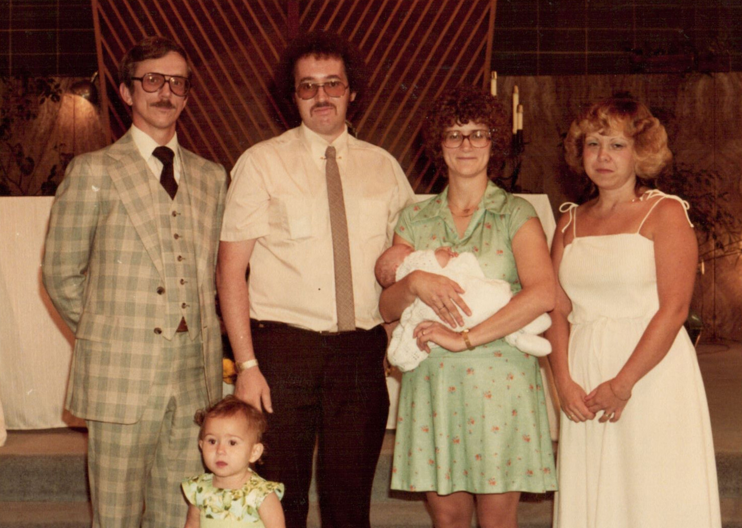 Family posing together indoors: two men (one in a plaid suit) and two women stand with a baby held by the woman in a green dress, plus a young girl in front.