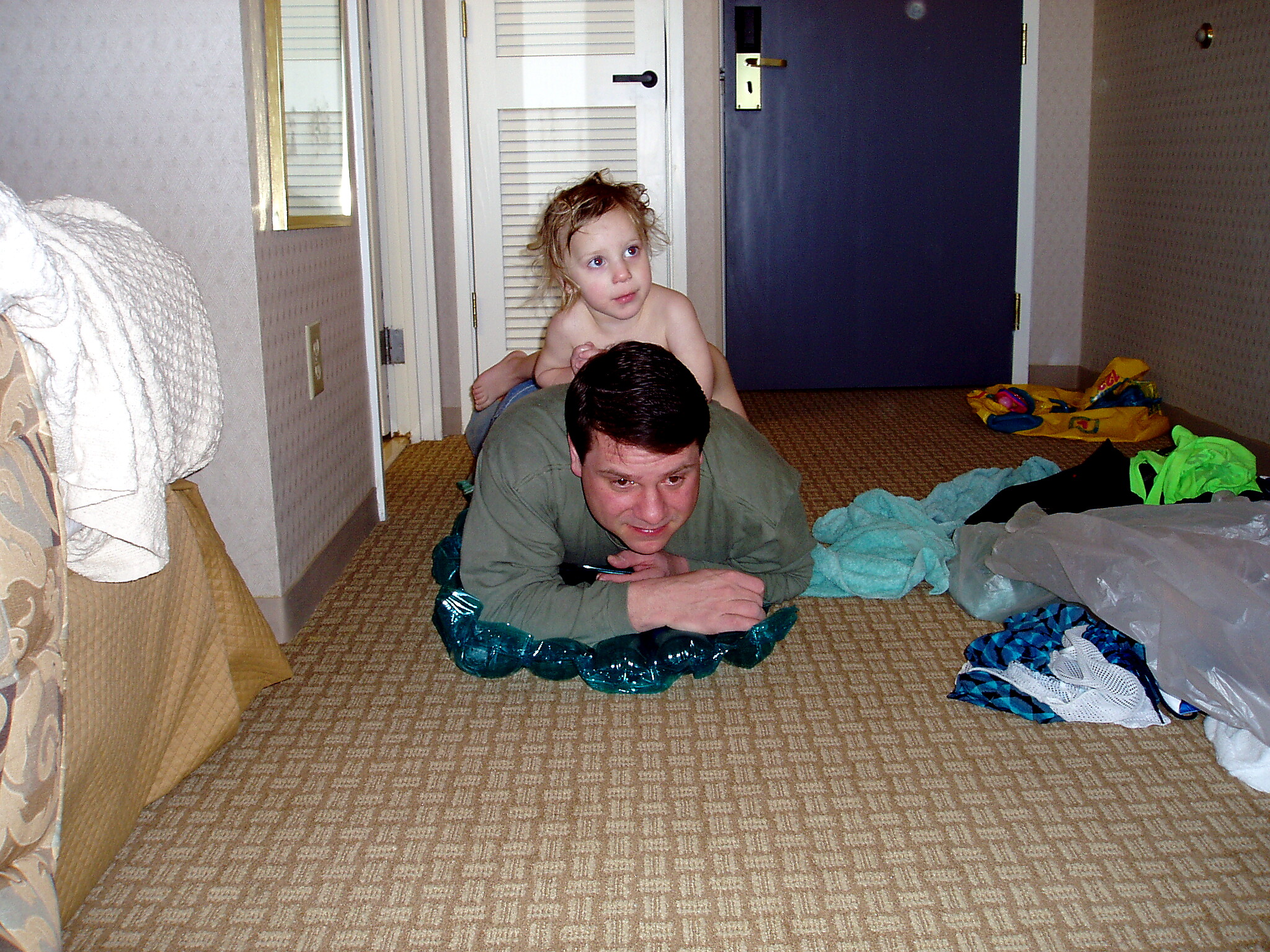Man on his hands and knees on a patterned hotel carpet, a small child sits on his back in a hallway with clothes strewn nearby