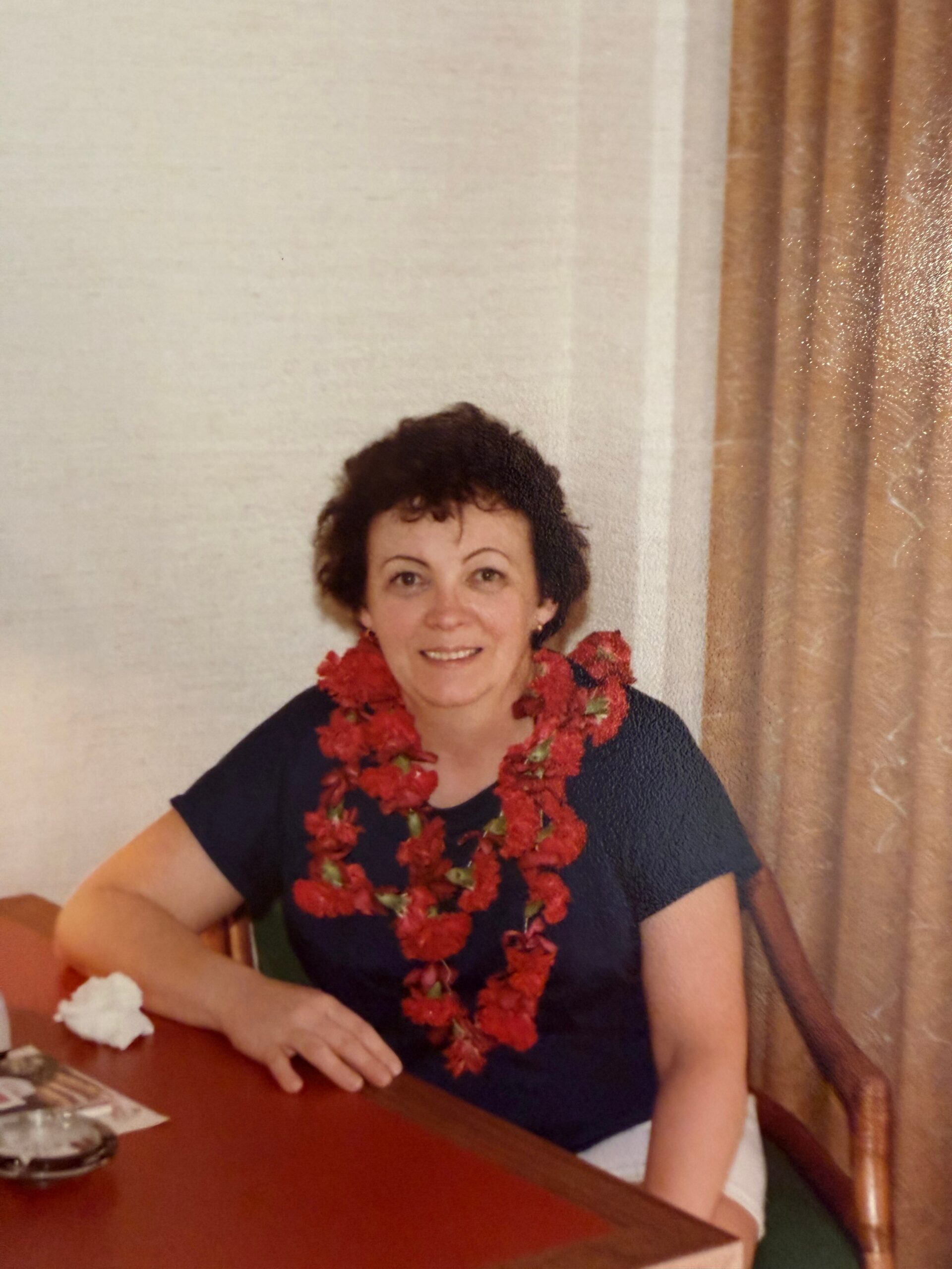 Smiling woman wearing a red flower lei, seated at a table indoors with beige curtains in the background.