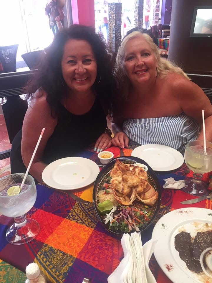 Two women smile at a restaurant table with a colorful tablecloth, sharing a plate of nachos and drinks. One wears a black top, the other an off‑the‑shoulder striped top.