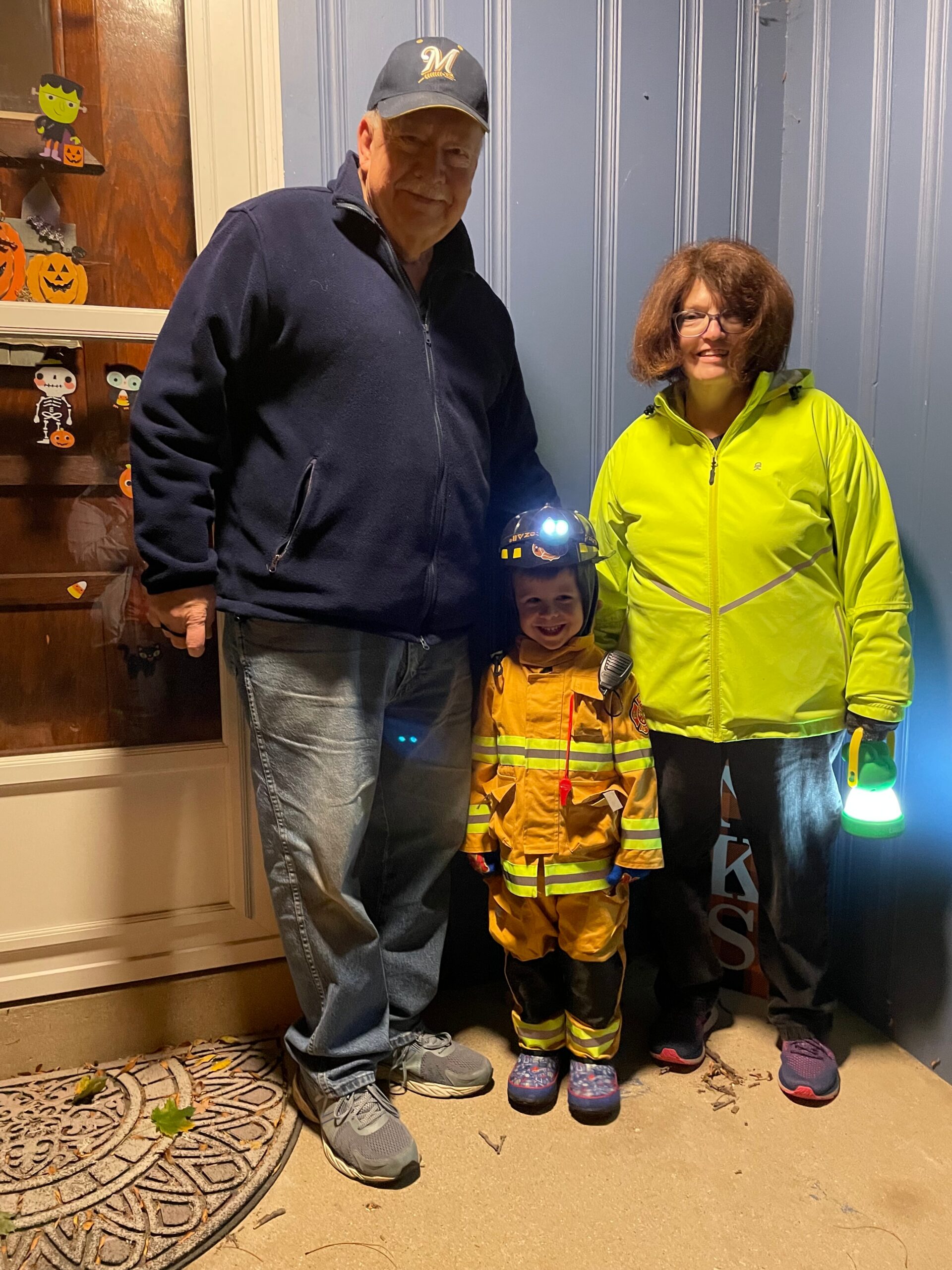 Three people pose together at a Halloween-decorated doorway: grandfather in a navy jacket and cap, a child in a firefighter costume with helmet, and a woman in a bright yellow jacket smile at the camera.