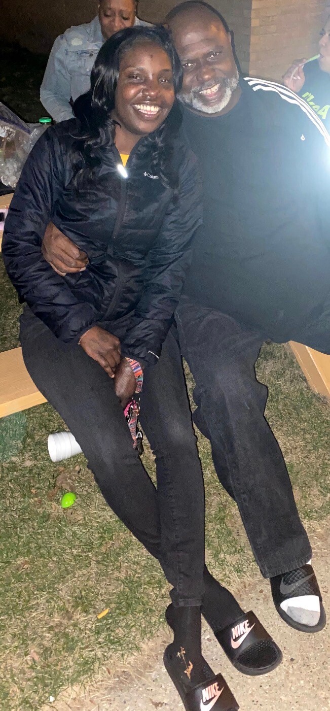 A smiling couple sitting close together on a bench outdoors at night, arms around each other, wearing jackets and casual shoes.