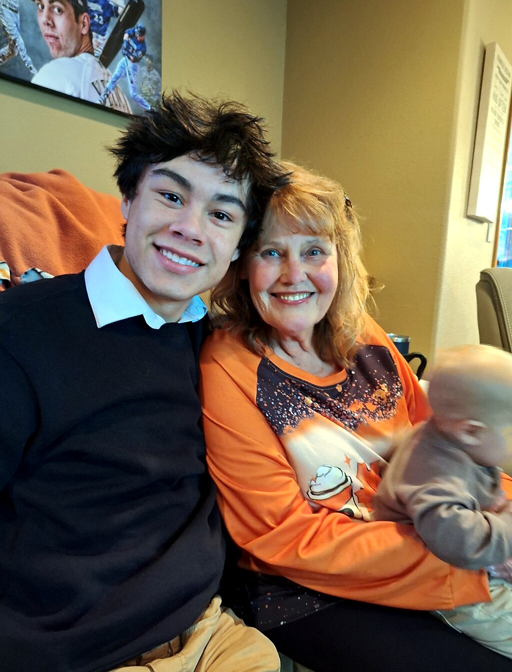 Smiling young man and older woman sit close together on a couch, with a baby on the woman's lap.