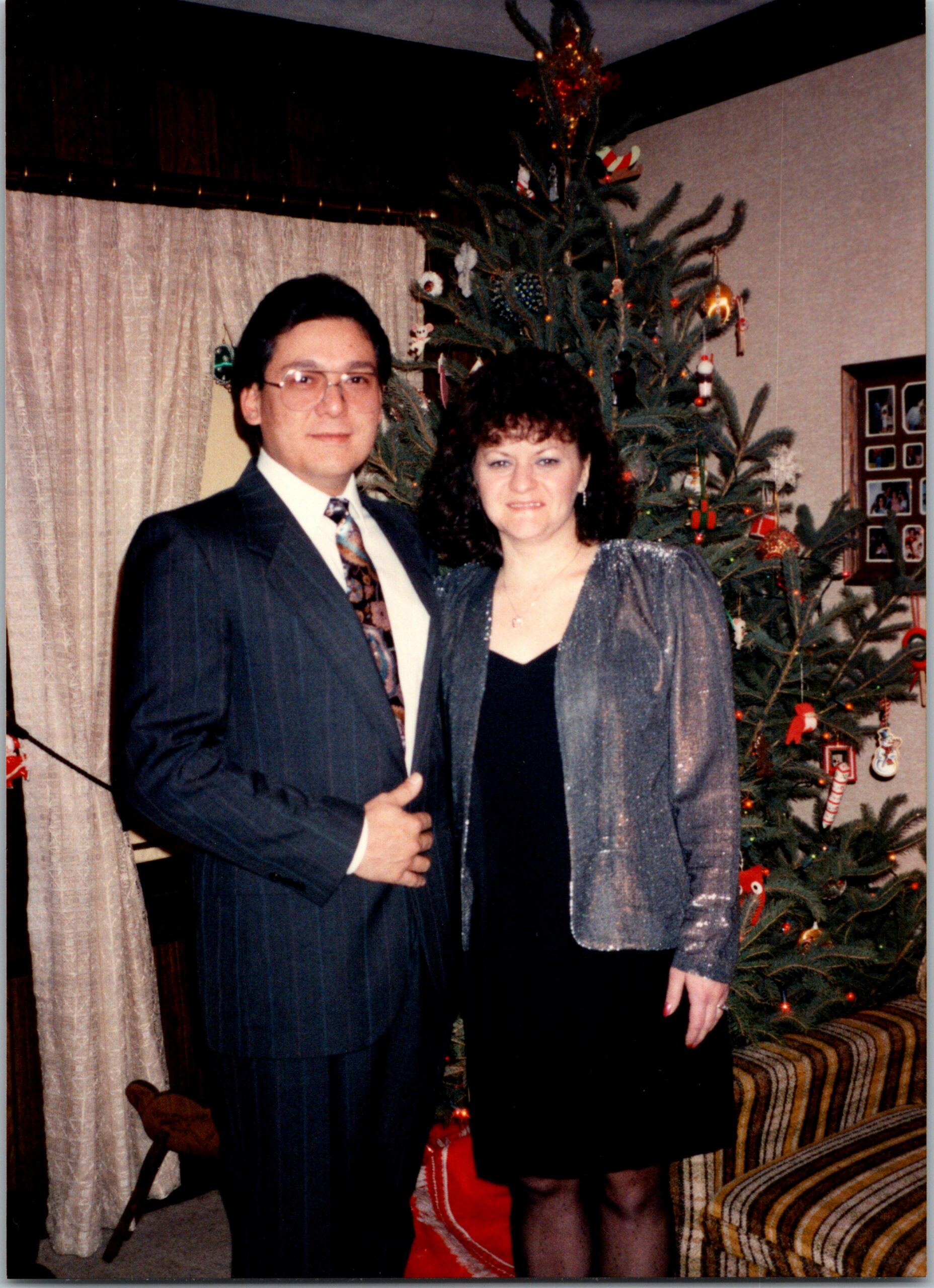 Couple posing by a decorated Christmas tree in a living room; man in a suit and woman in a sparkly jacket.