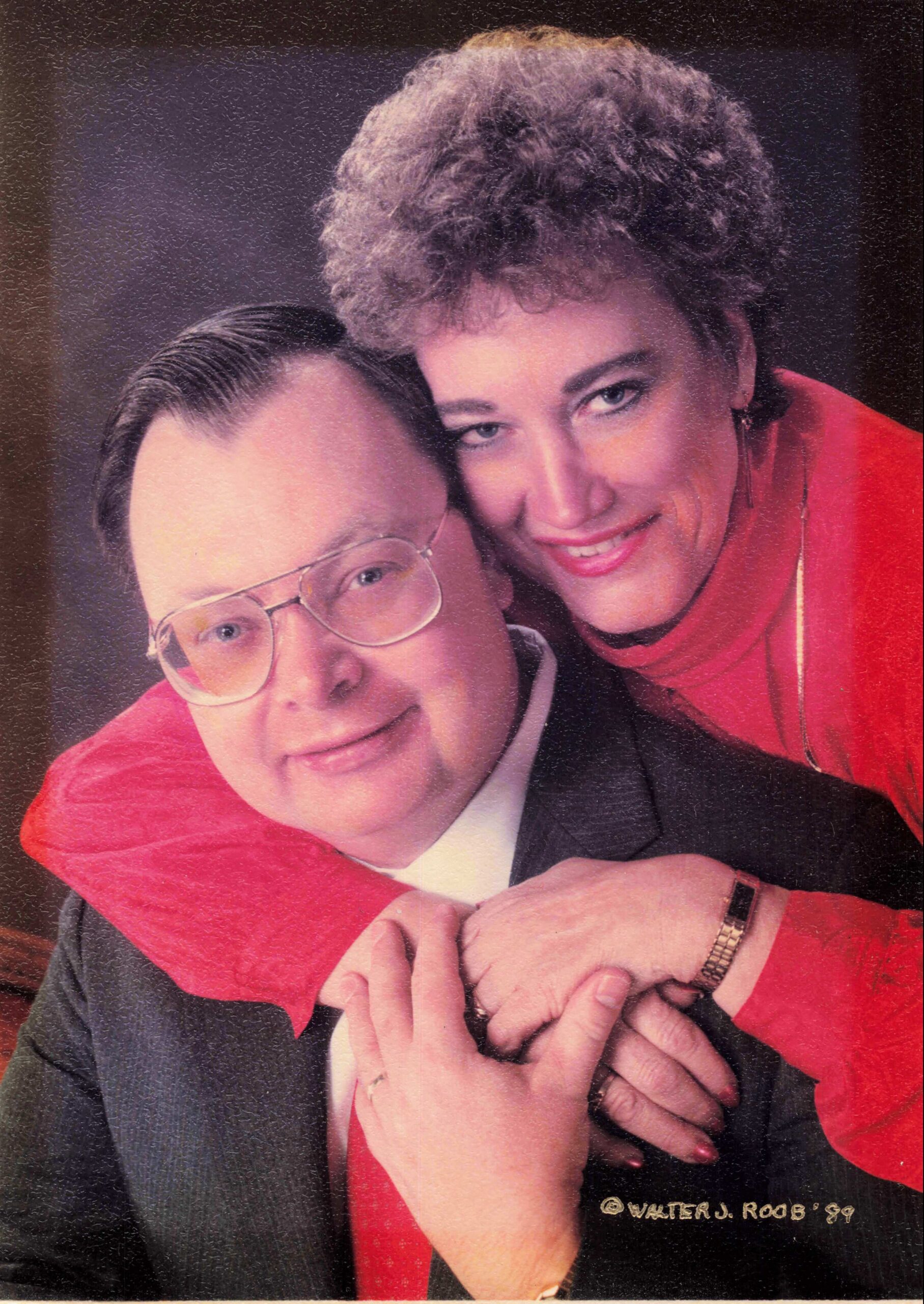 Couple hugs and smiles at the camera; man with glasses in a dark suit and a woman in a red outfit.